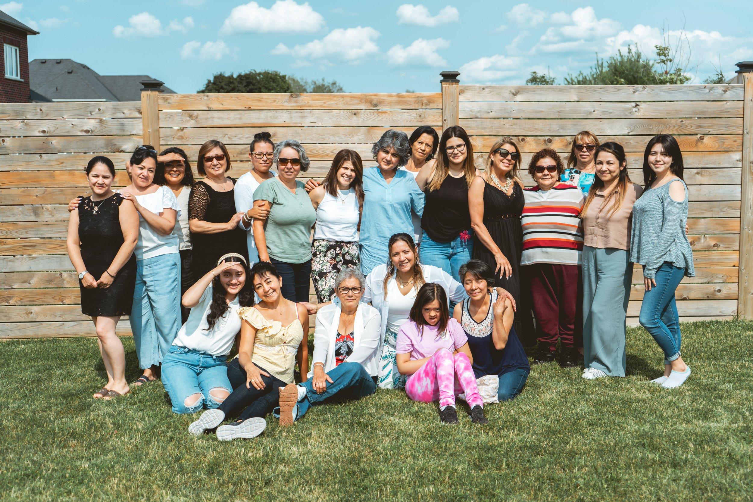 A group of women posing together outdoors in front of a wooden fence on a sunny day, standing and seated on grass.
