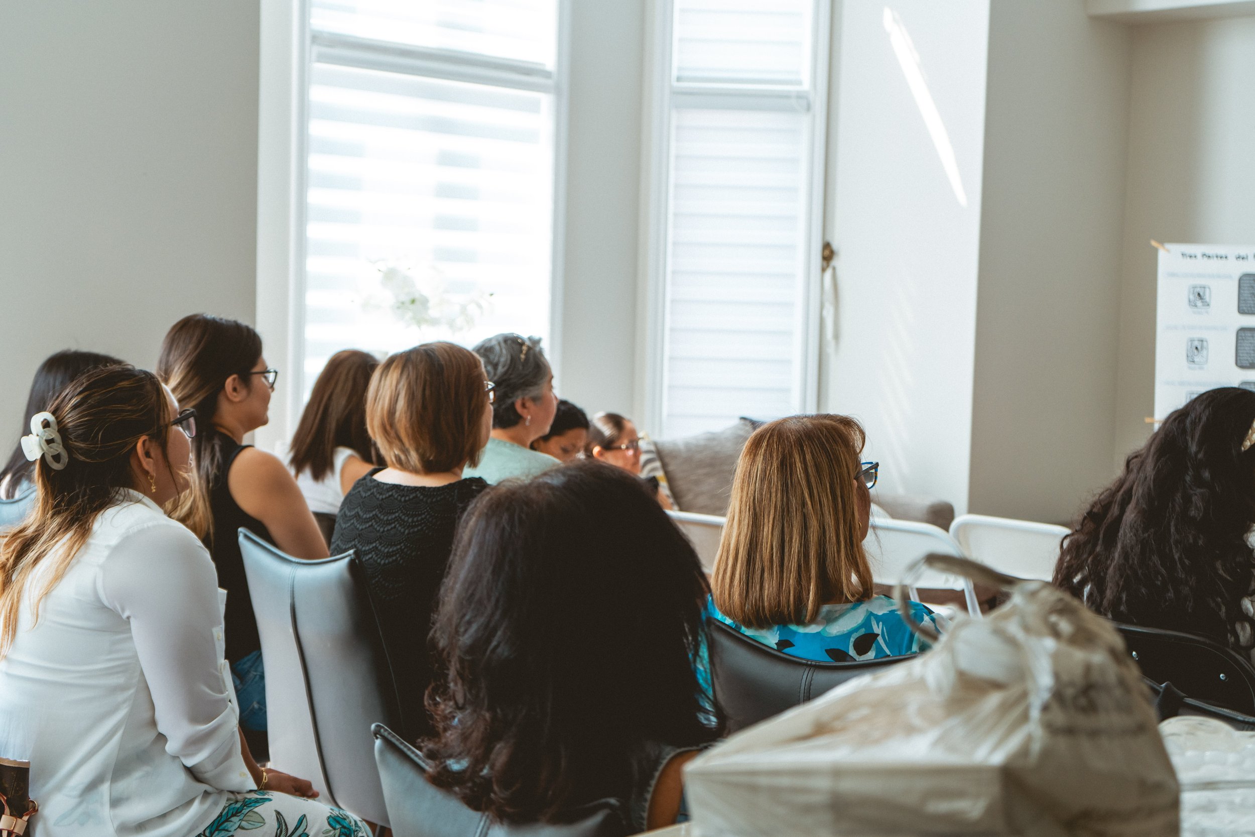 A group of people seated indoors, facing forward, watching a presentation or speaker. They are in a bright room with light coming through the window.