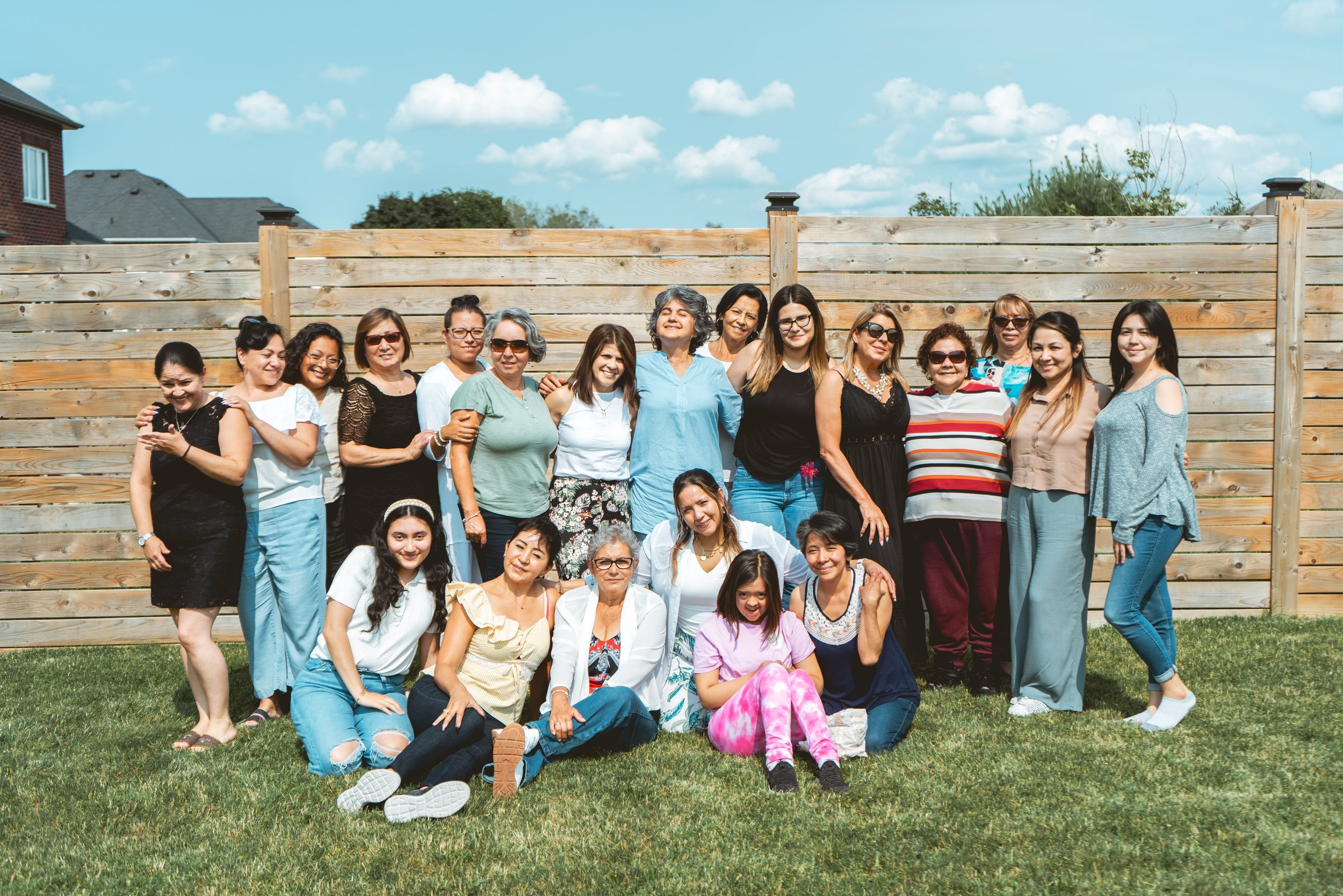 Group of women posing outdoors in front of a wooden fence on a grassy lawn.