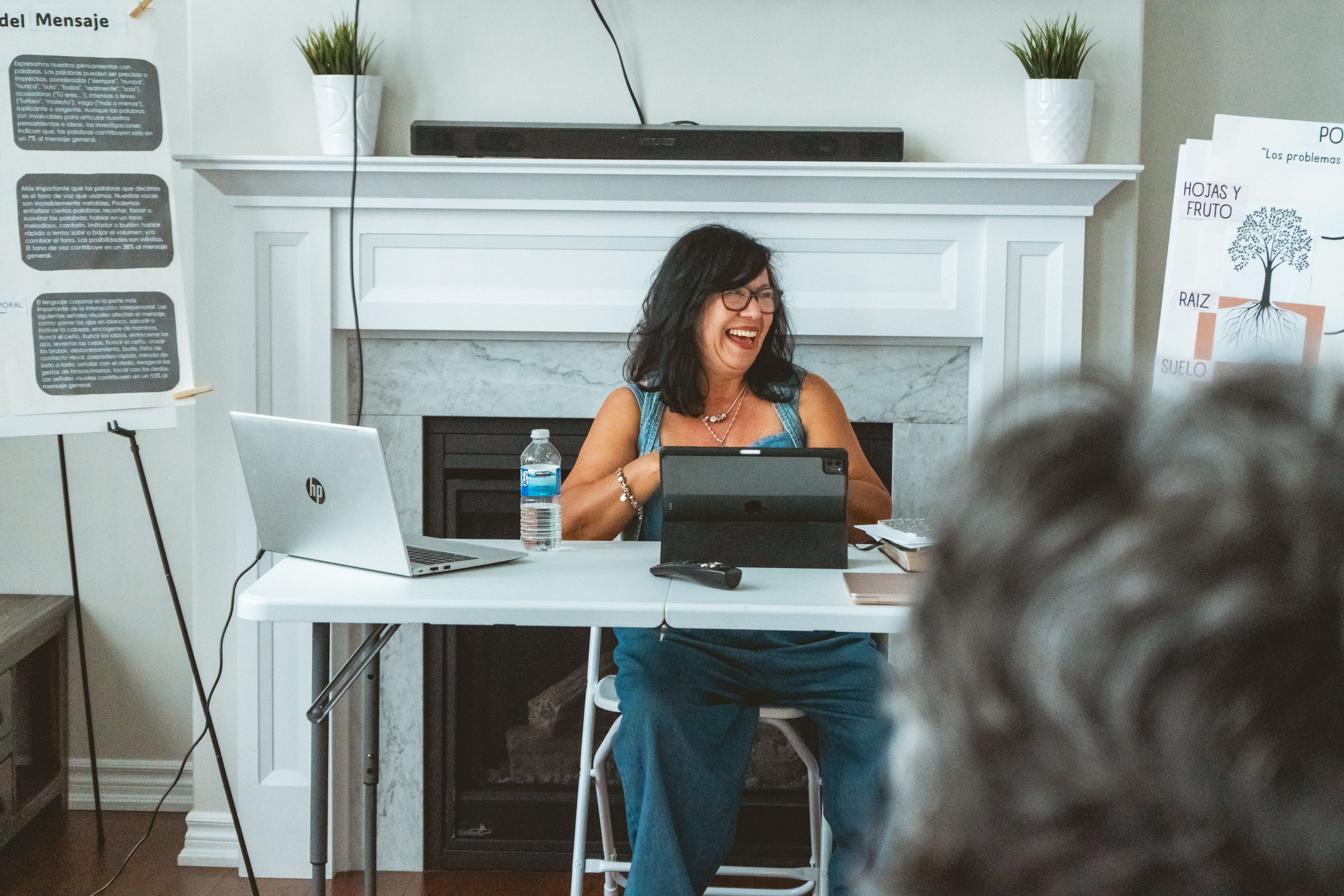 Woman sitting at a desk with a laptop and tablet, smiling during a presentation. A white fireplace mantle with potted plants is in the background, along with poster boards with text and diagrams.