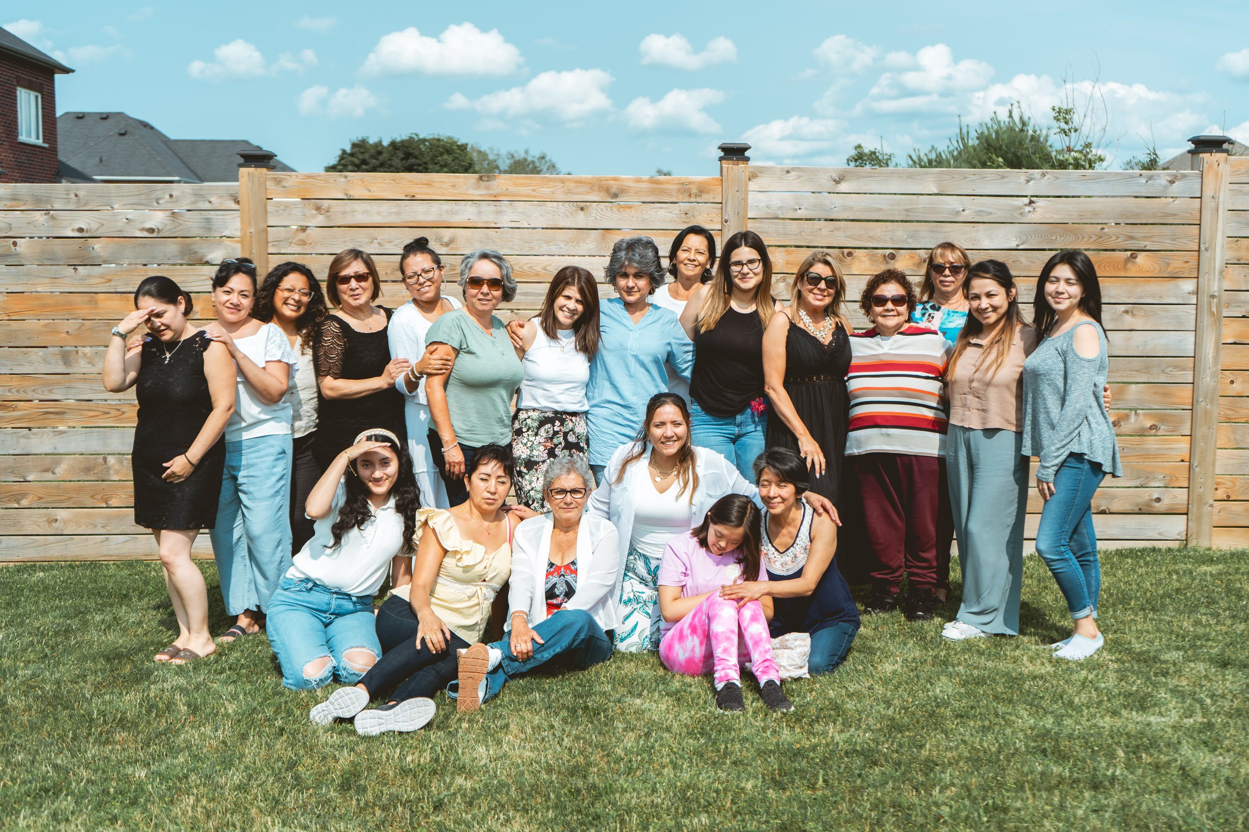 Group of women posing outdoors in front of a wooden fence on a sunny day, some sitting on grass.