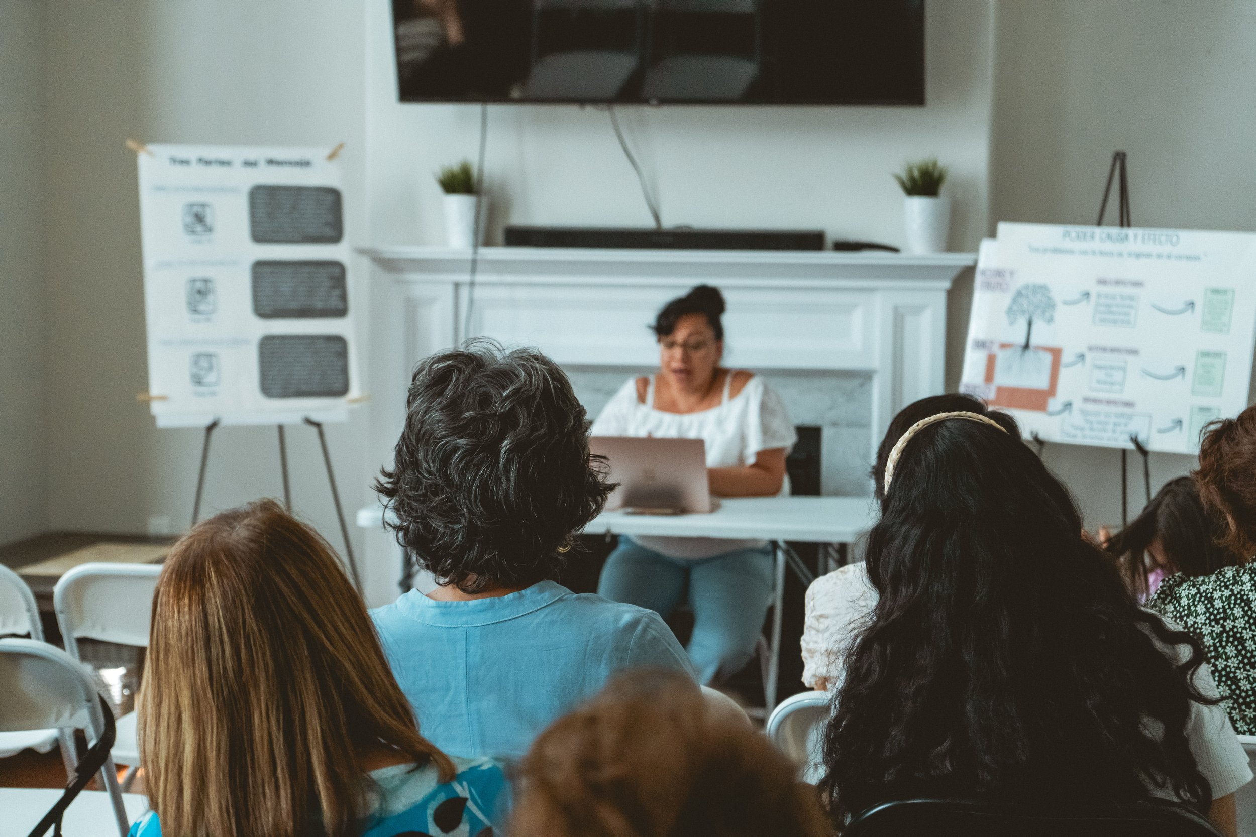 A woman sits at a table in front of an audience speaking or presenting. There are two easels with charts on either side of her. The setting appears to be a casual meeting or workshop. The audience's backs are visible, indicating they are listening at