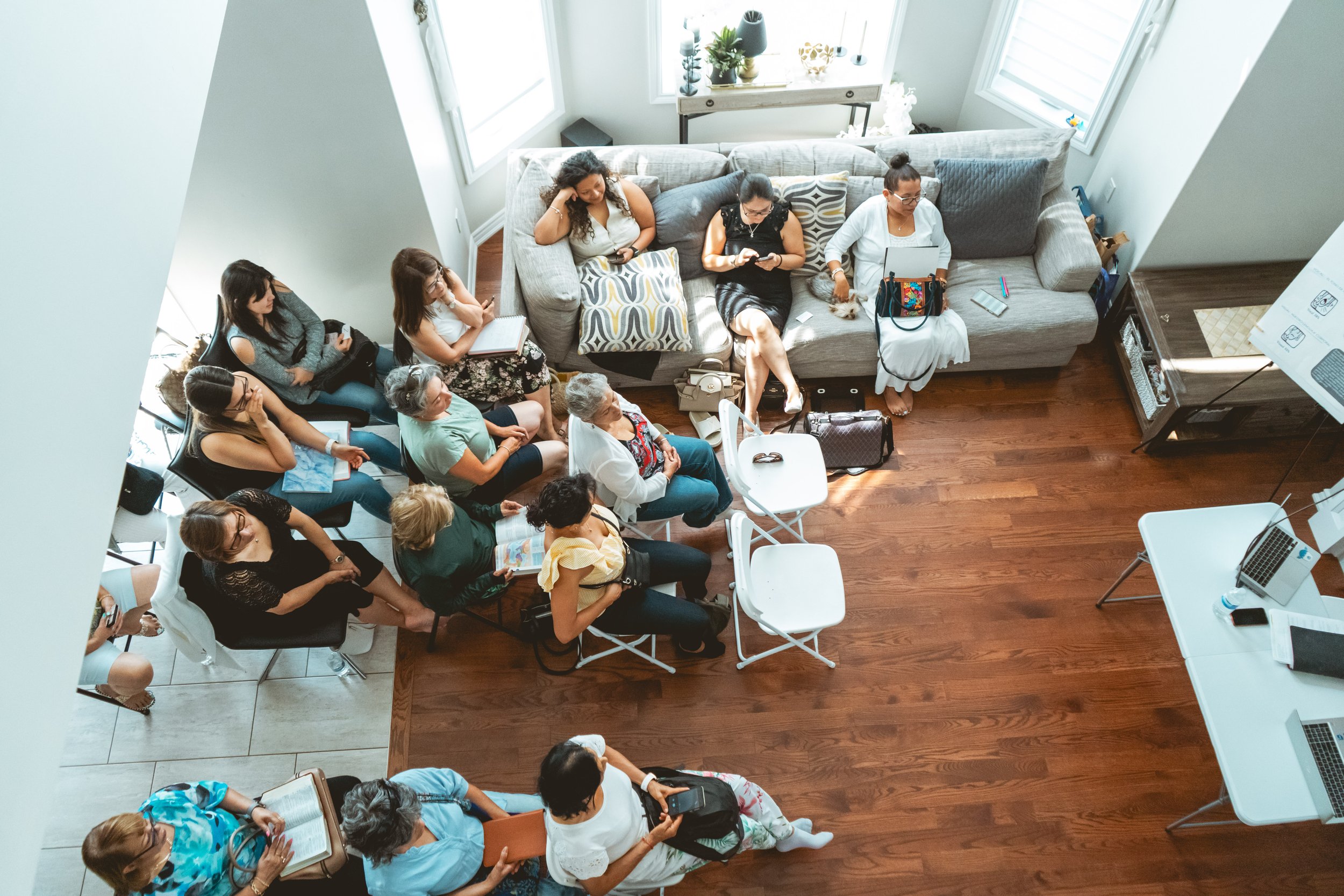 Overhead view of a casual indoor gathering, with people sitting on a sofa and folding chairs, some using laptops and smartphones, in a well-lit room.