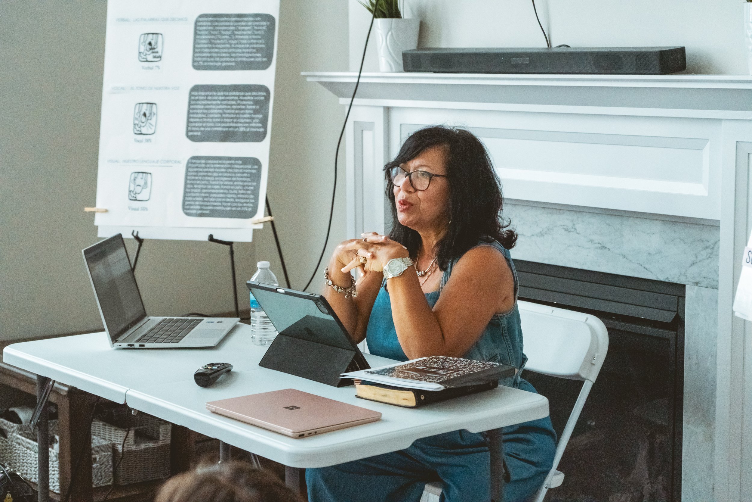 A woman sitting at a table giving a presentation with laptops, a tablet, a notebook, and a water bottle. A poster with diagrams and text is in the background.
