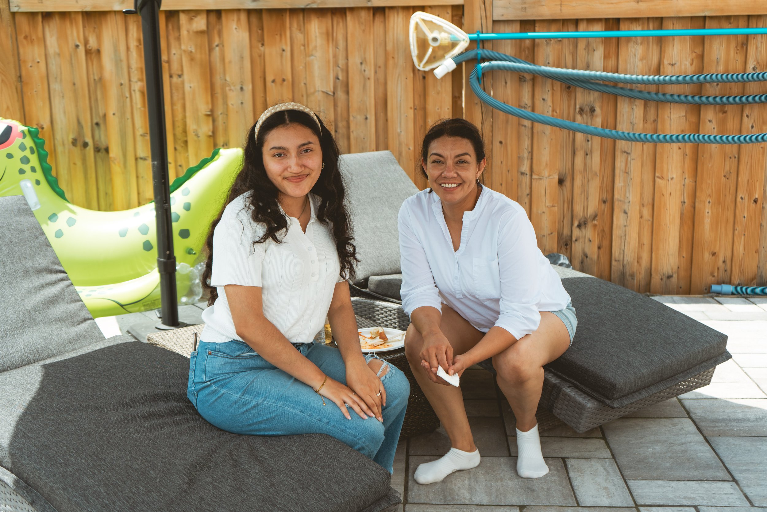 Two women sitting on lounge chairs by a pool with a wooden fence and inflatable pool toy in the background.