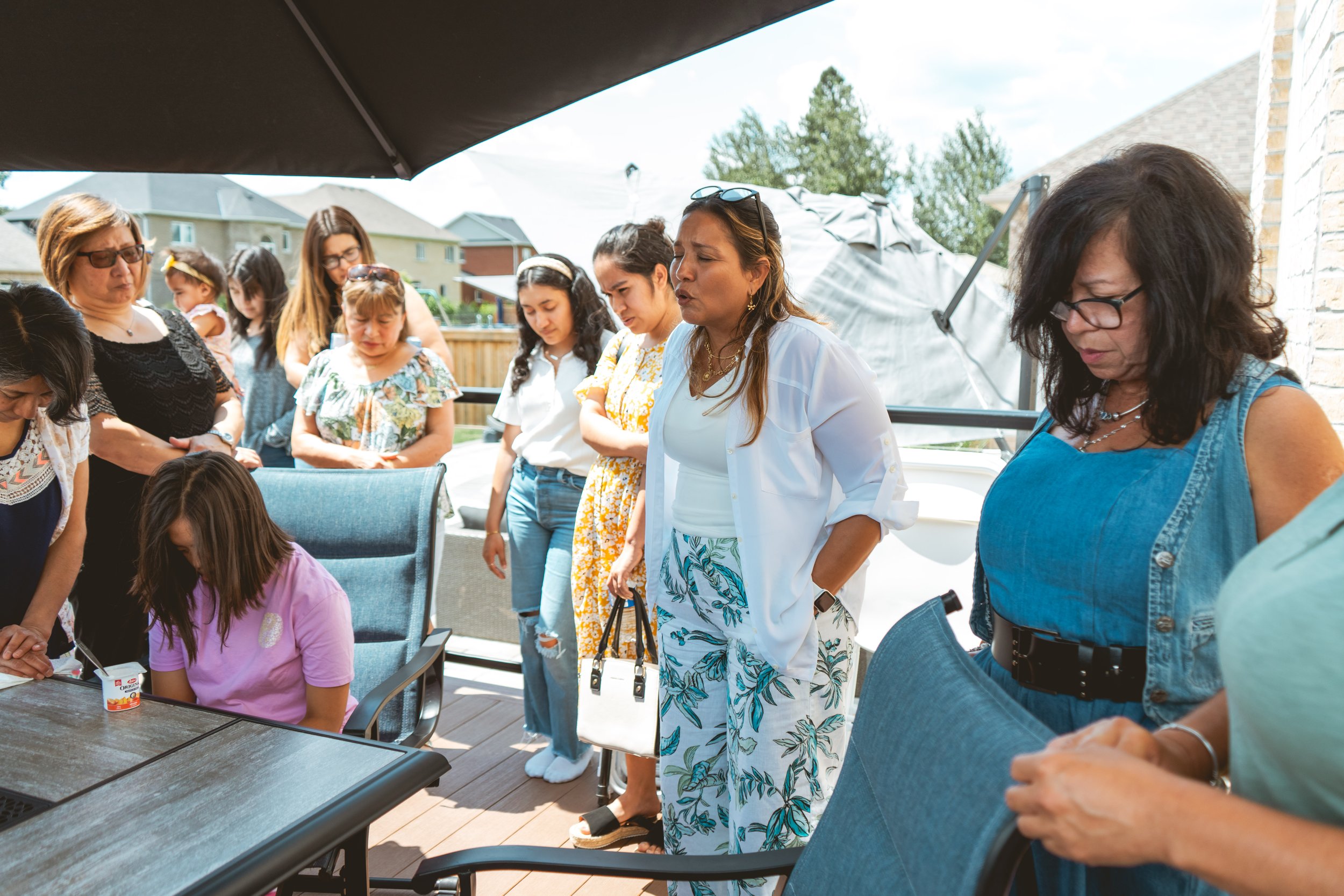 A group of women and a child gathered outdoors around a table. One woman is seated while others stand, some looking down. The setting is a residential backyard with patio furniture and houses in the background.
