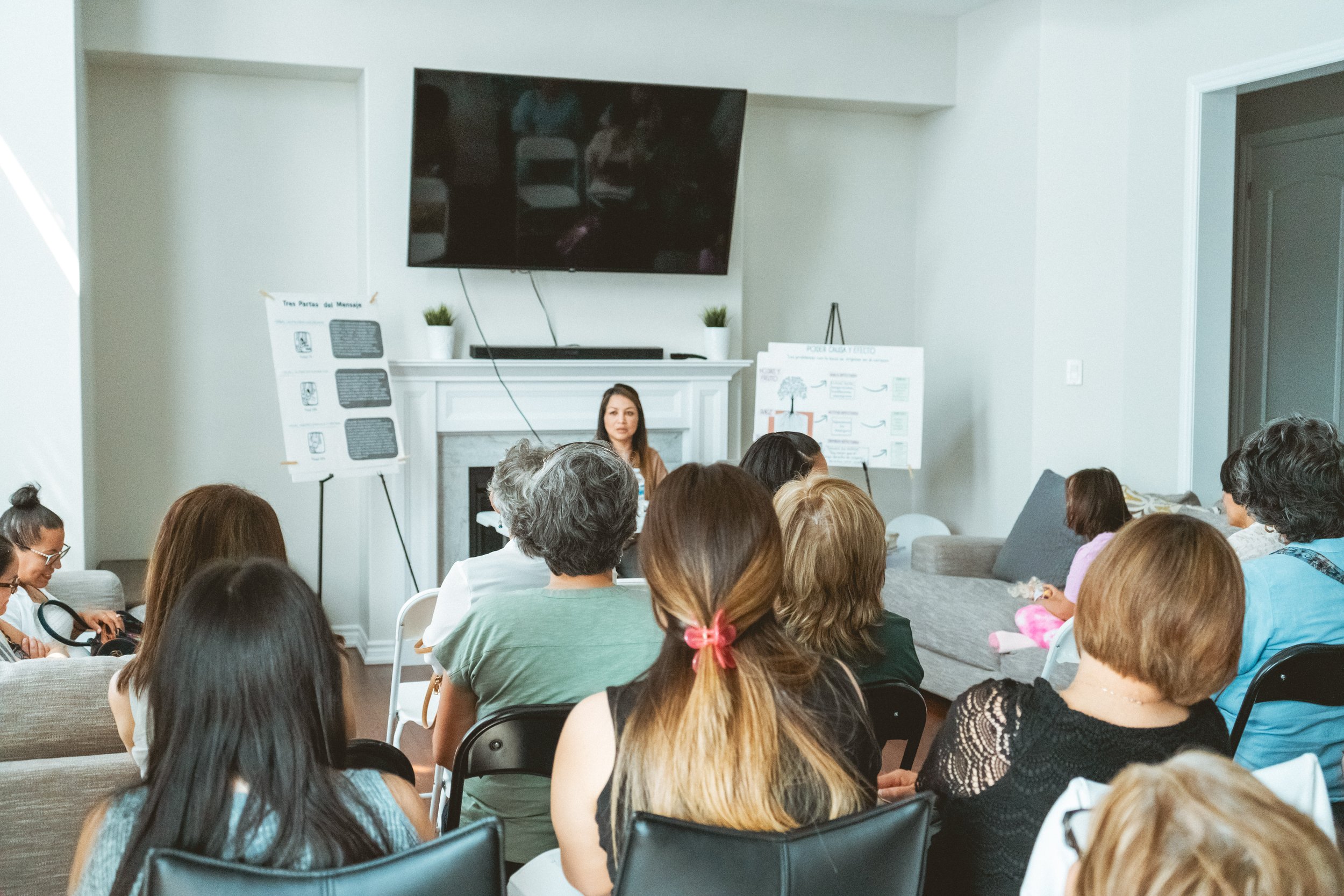 A woman speaking to a group in a living room, with presentation boards and a large TV screen in the background.