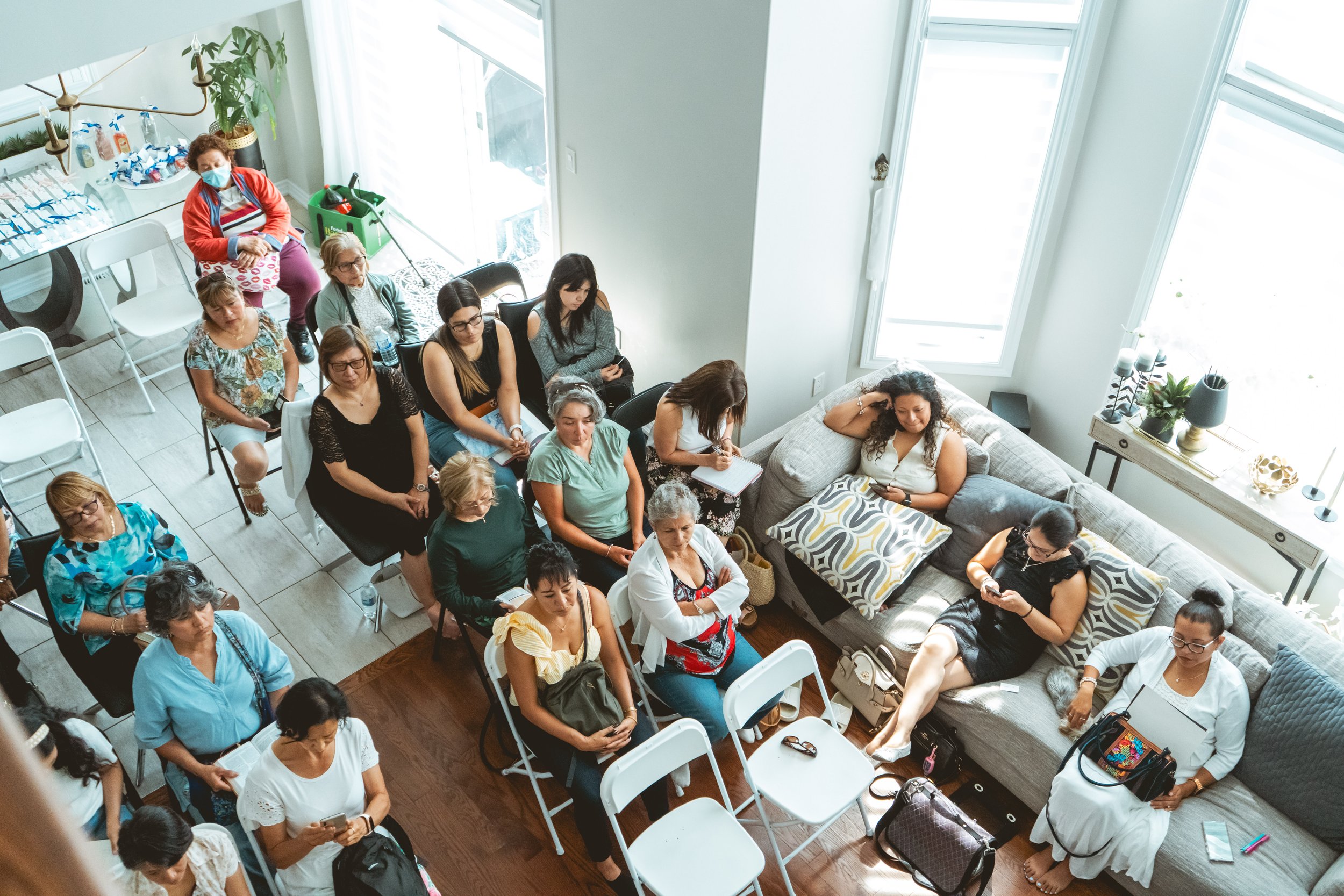 A group of women seated indoors, some on a couch and others on folding chairs, in a bright room with large windows. Some are using mobile devices, while others are listening attentively. The background includes a table with refreshments and a few ind