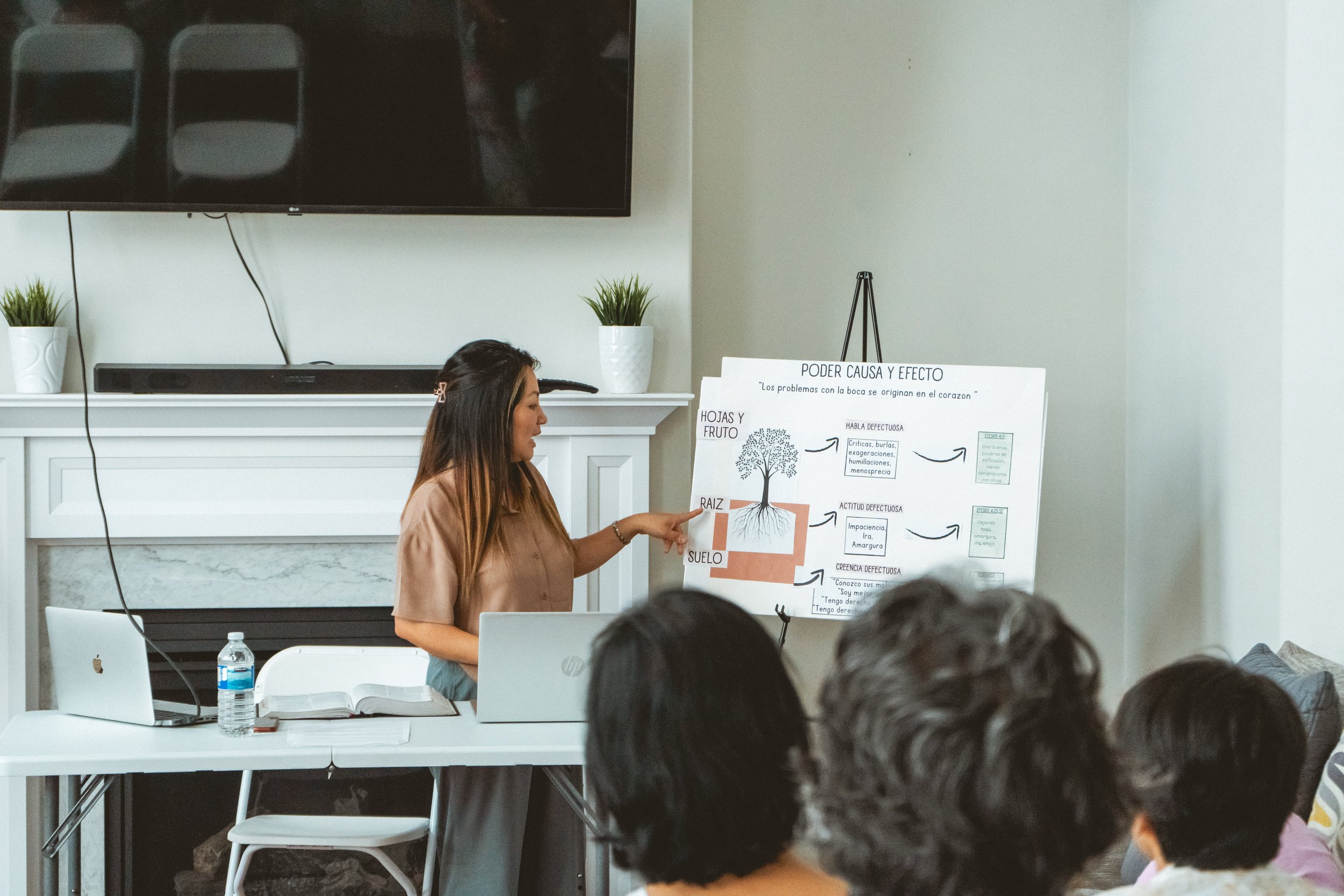 A woman presenting a diagram at a seminar, pointing at a poster on an easel, with a table holding laptops and a water bottle in the foreground. Participants sit facing her.