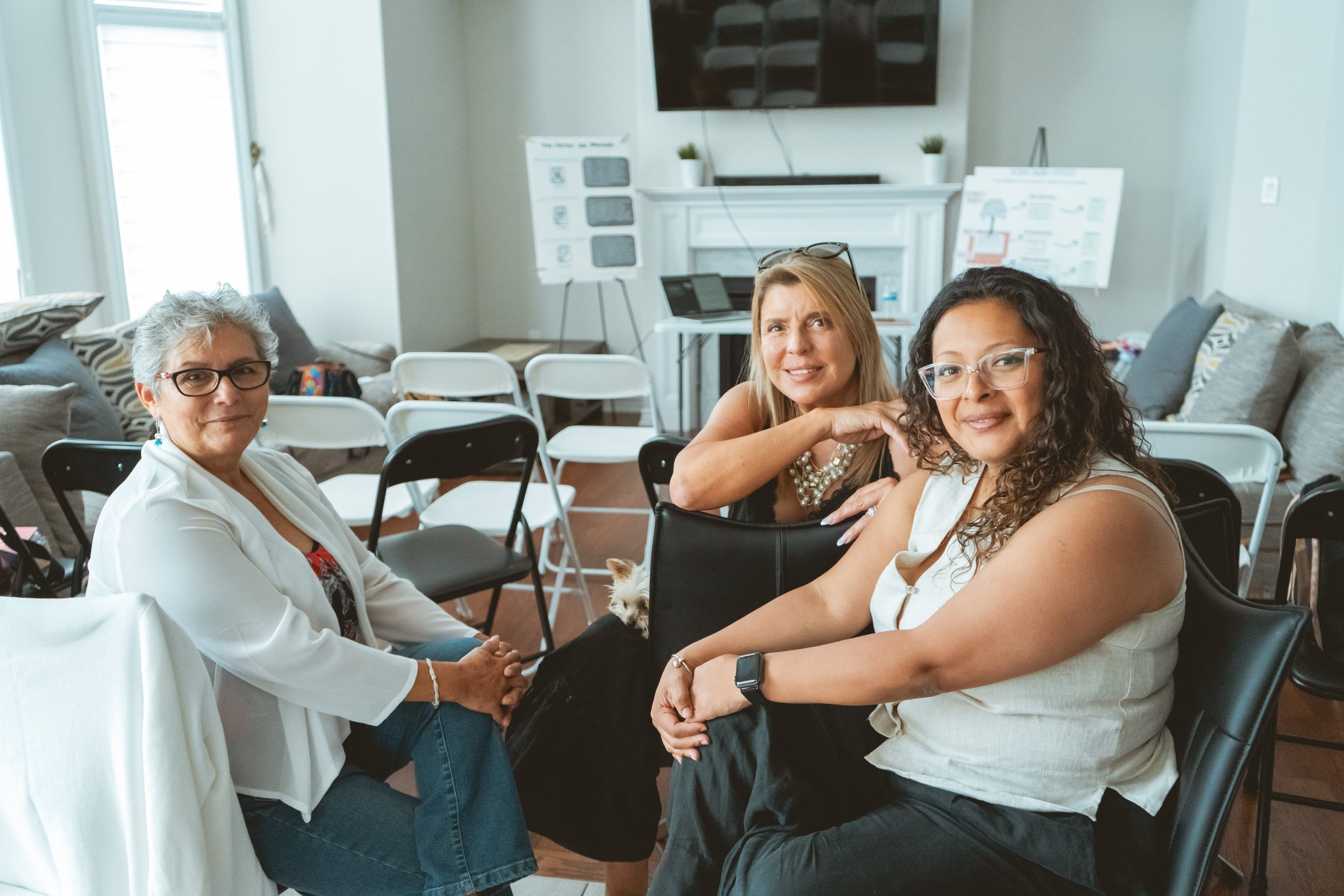 Three women sitting in a room with chairs and presentation boards in the background.