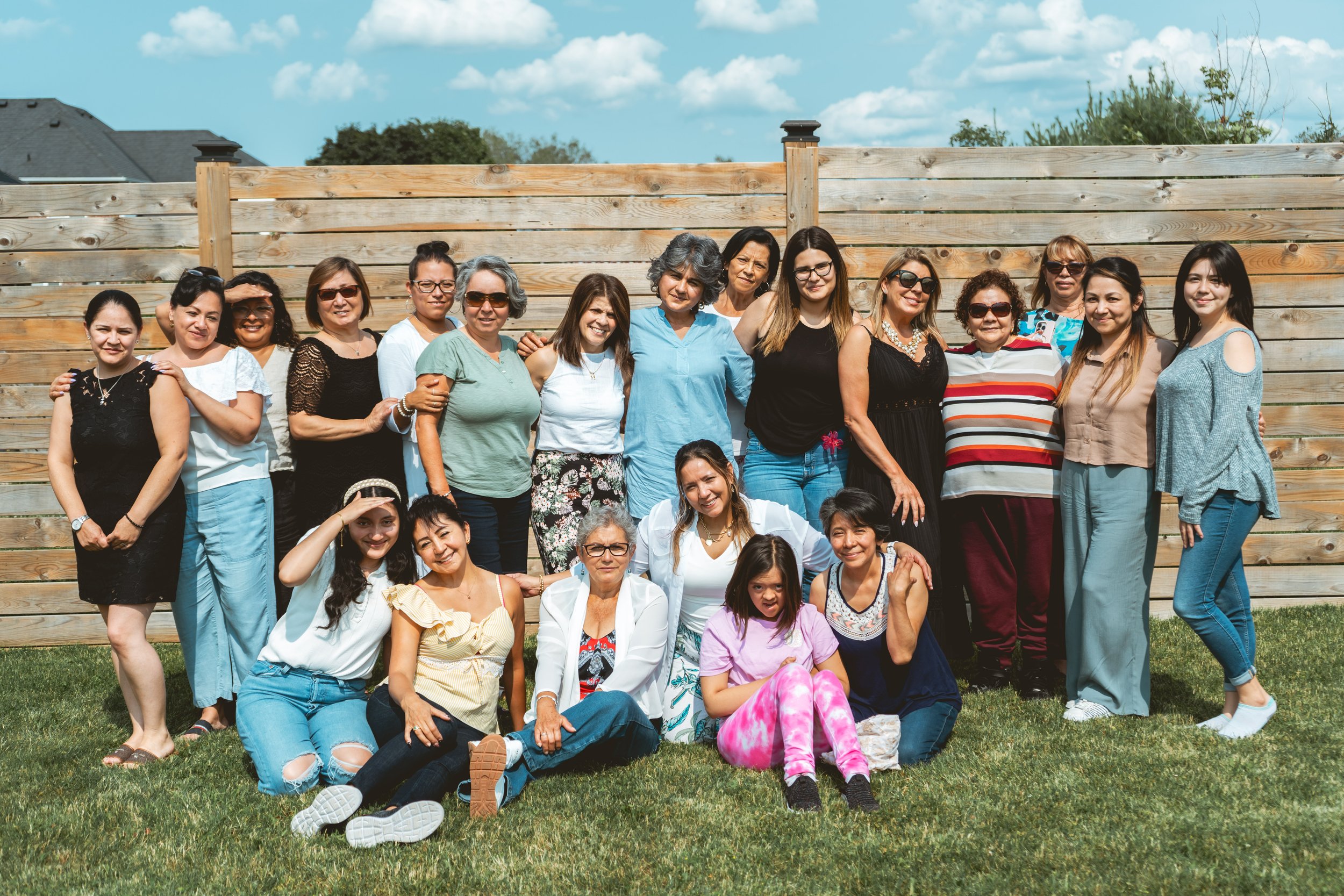 Group of women posing outdoors in front of a wooden fence under a blue sky.