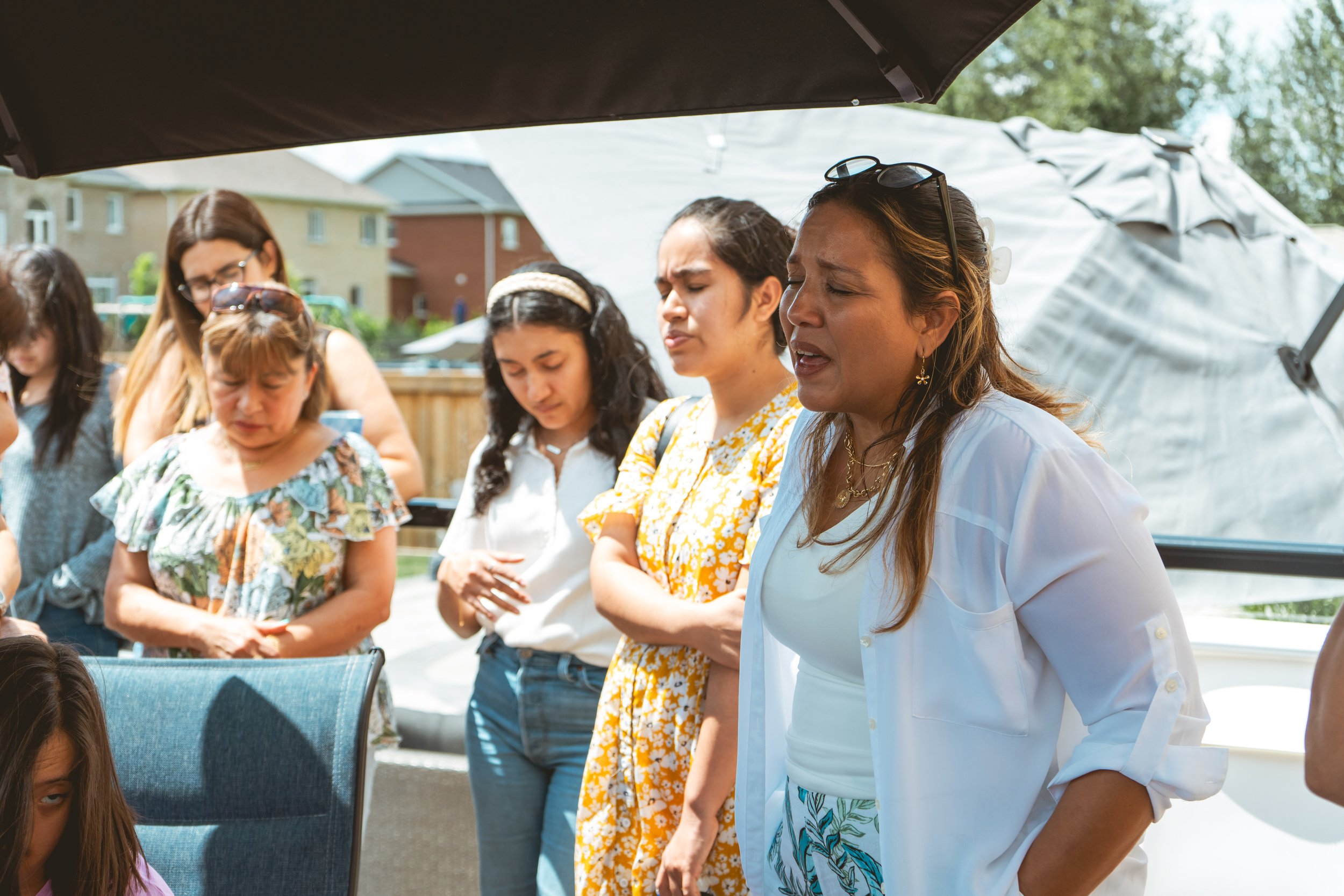 A group of people standing outside, some looking down or with eyes closed, possibly in prayer or deep thought. The setting is in a backyard with a fence and houses visible in the background. The weather appears sunny.