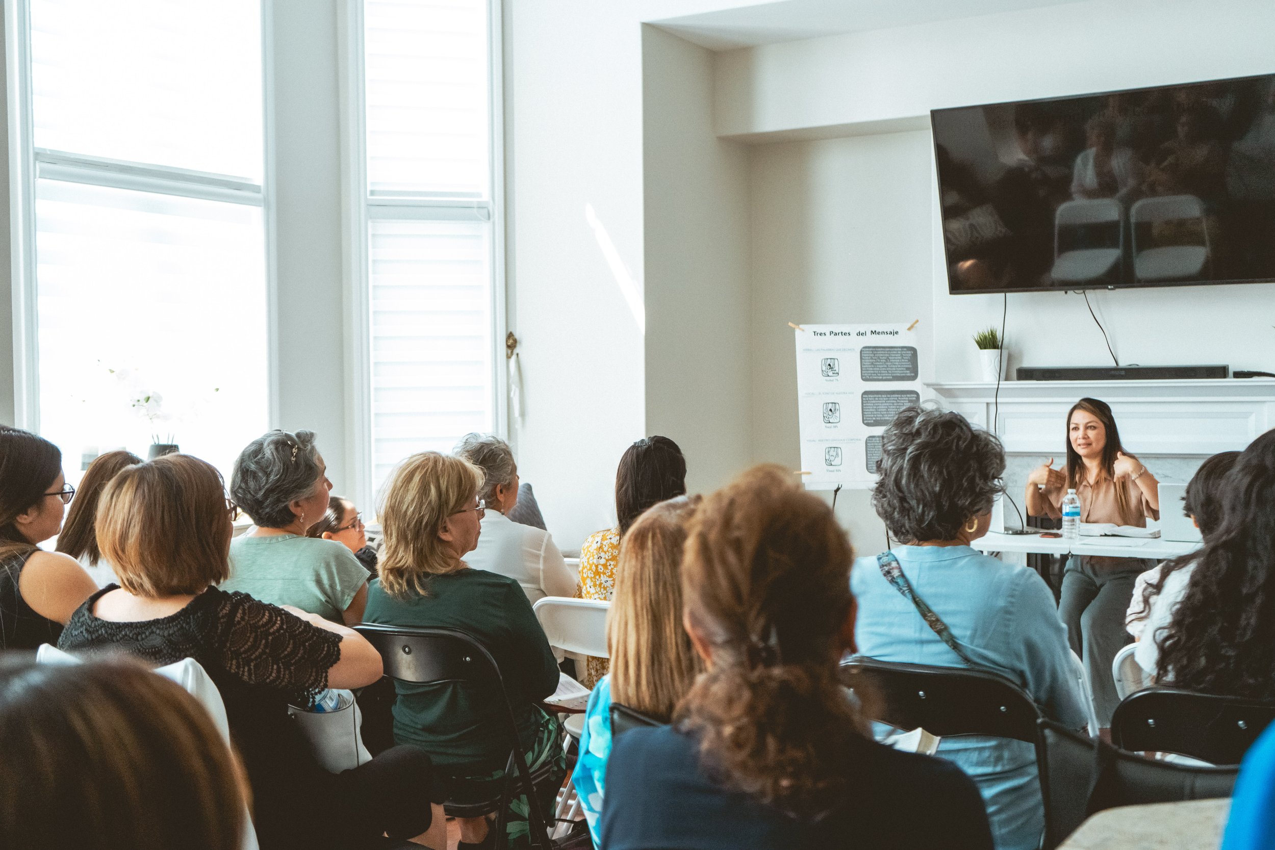 A woman speaking to a group of people in a seminar setting with a paper titled "Tres Partes del Mensaje" on display.