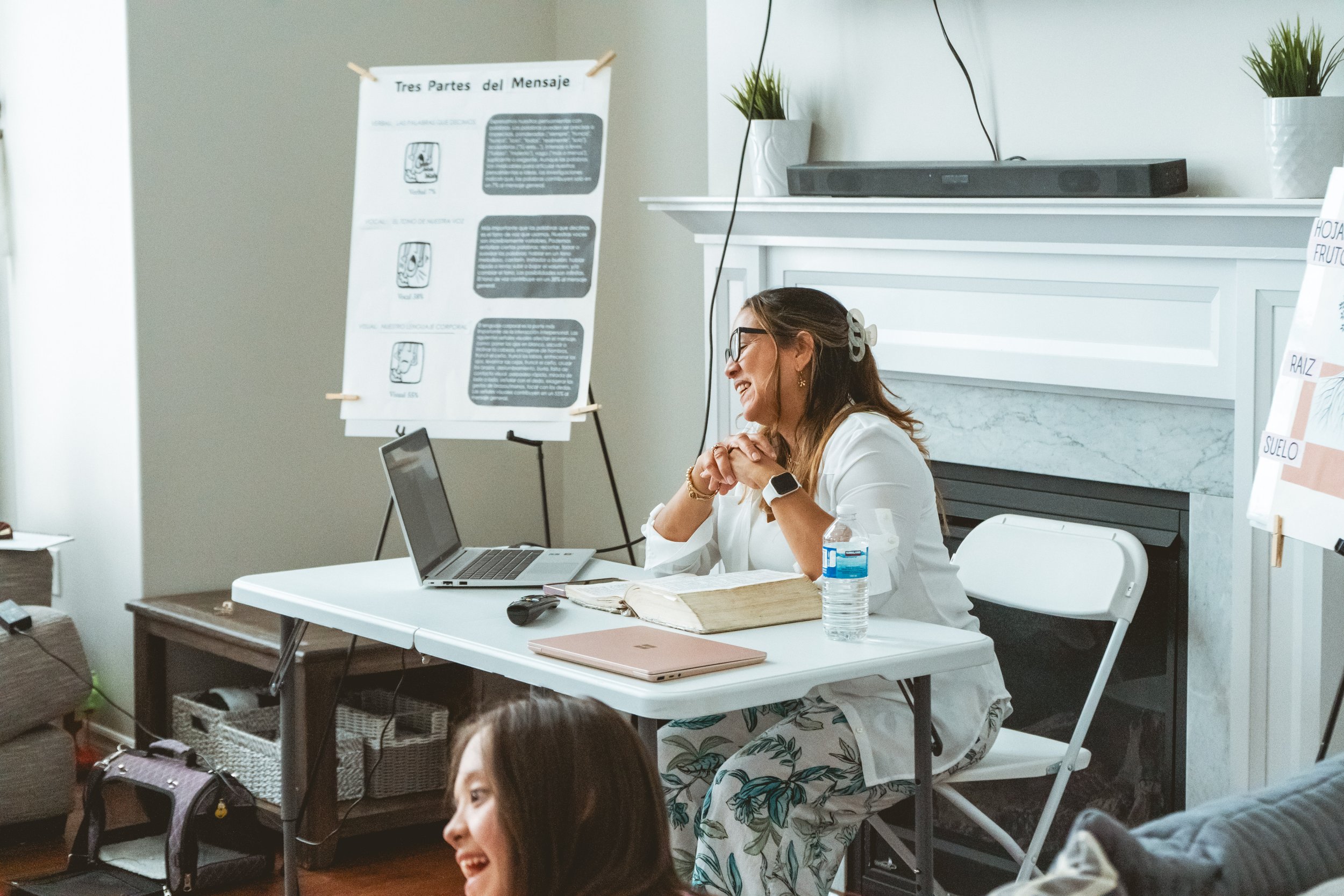 A woman is sitting at a table with a laptop, book, and water bottle, engaging in a discussion. There's a chart with "Tres Partes del Mensaje" in Spanish on a stand, and a whiteboard in the background with notes.