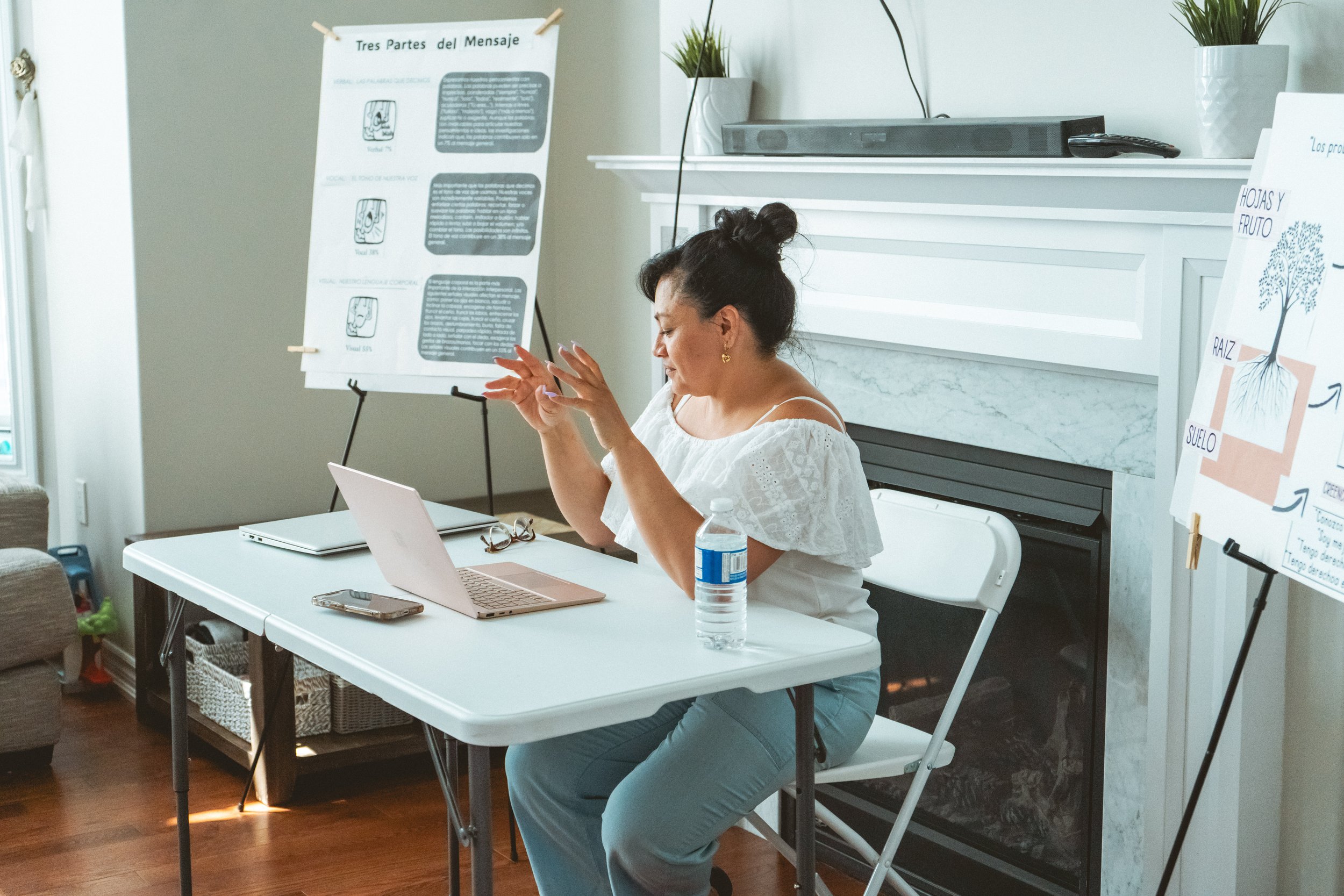 A woman gestures while sitting at a table with a laptop, a smartphone, glasses, and a water bottle. Behind her, a whiteboard with Spanish text is visible, next to a fireplace and two plant pots on the mantel.