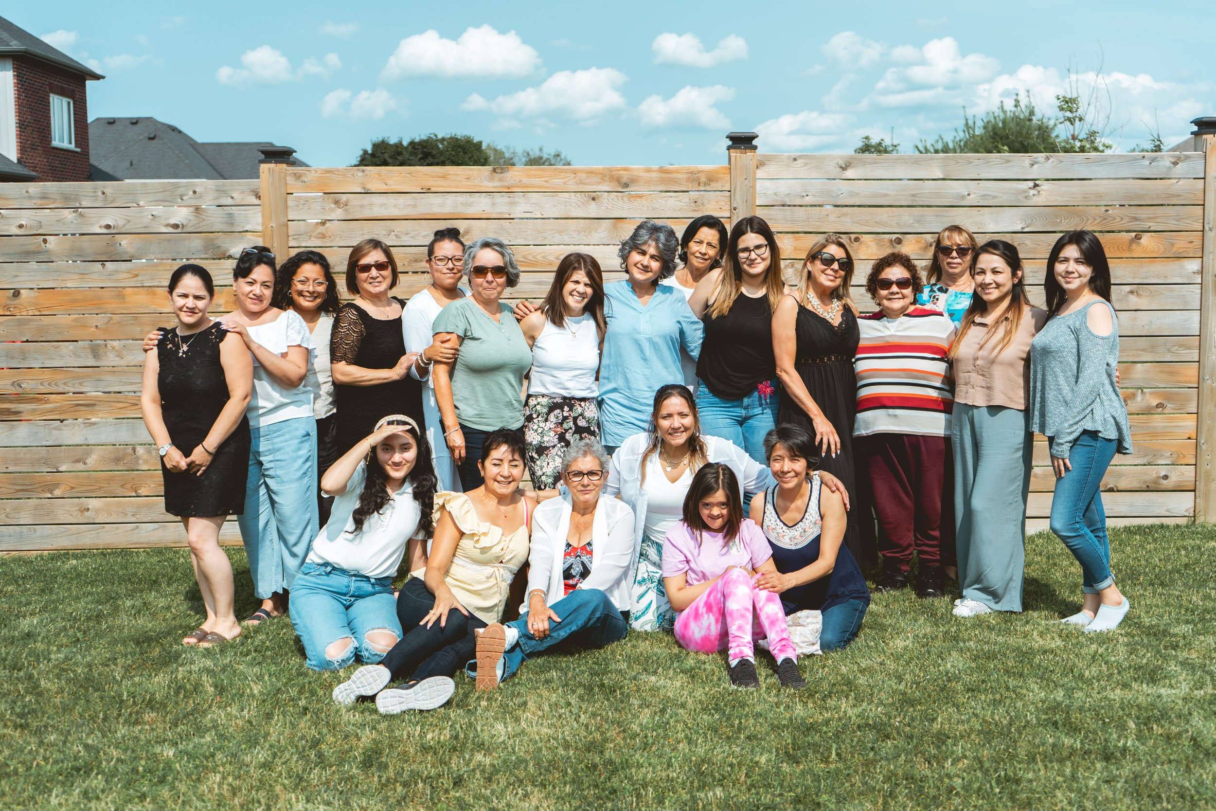 Group of women and children posing outdoors in front of a wooden fence on a sunny day.