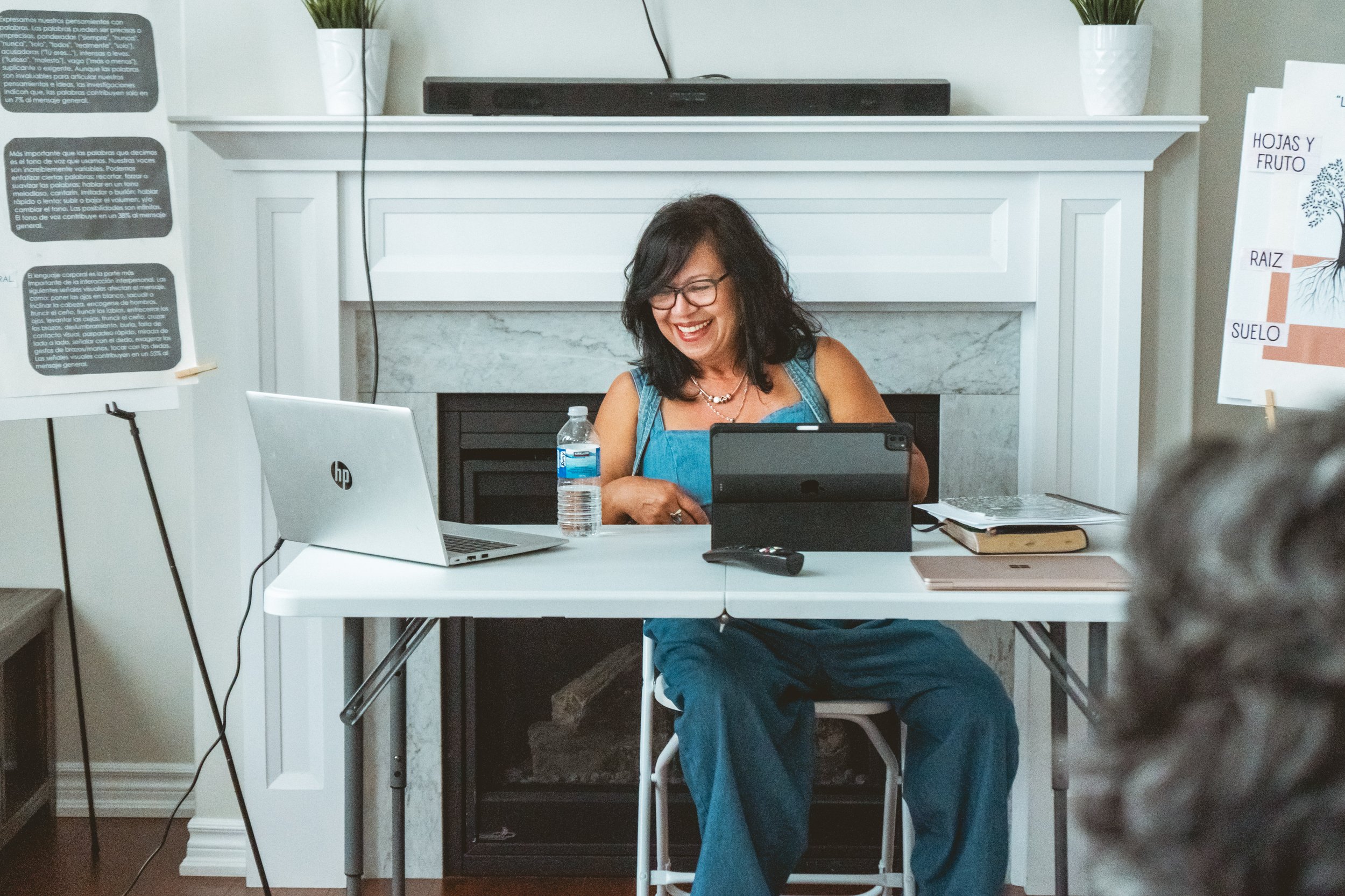 Woman sitting at a table in front of a fireplace, using electronic devices, with a water bottle and books. Presentation materials are on easels nearby.