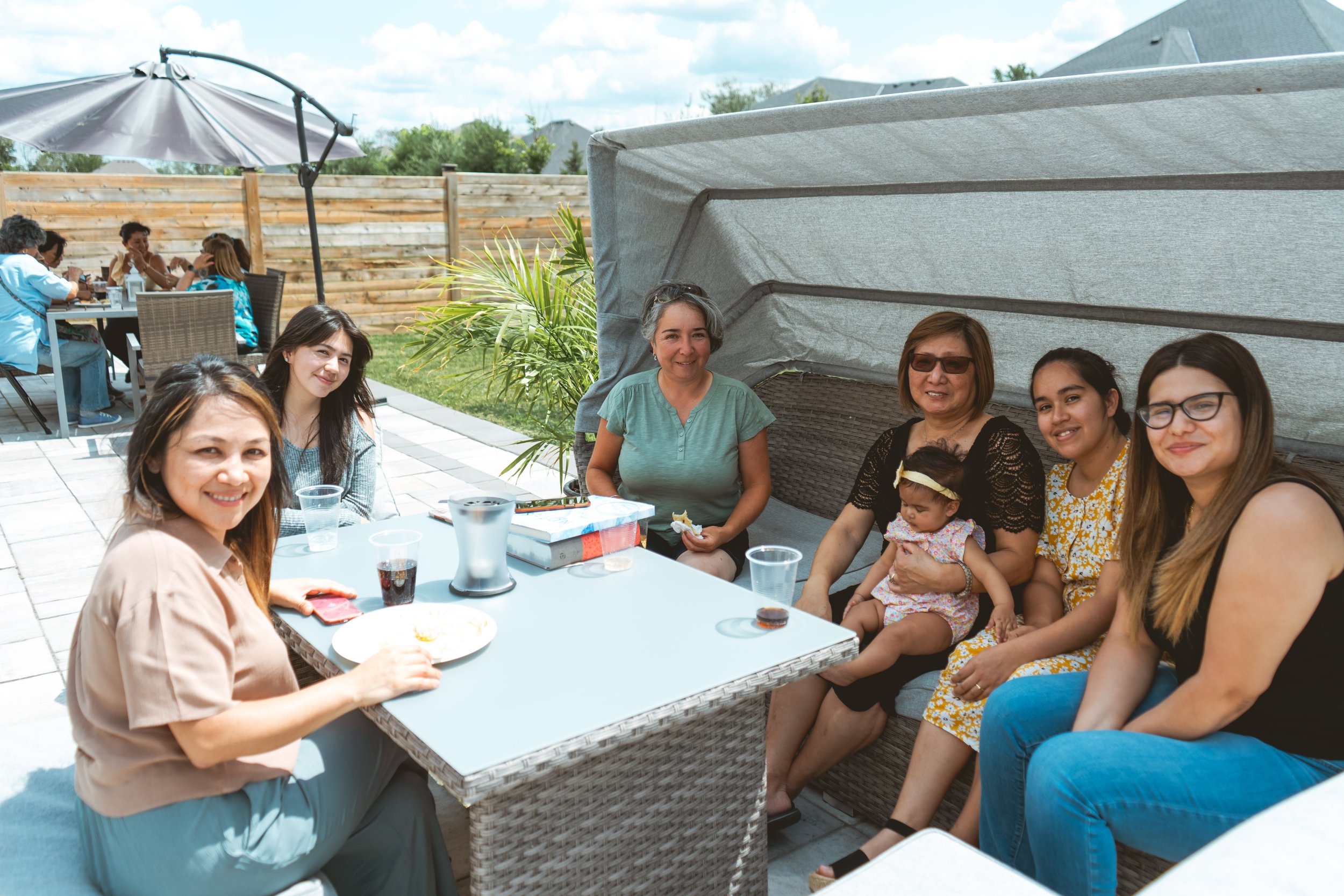 A group of women and a baby sitting together outdoors on a patio, enjoying a meal and drinks around a table. The setting is sunny and they are under a canopy.
