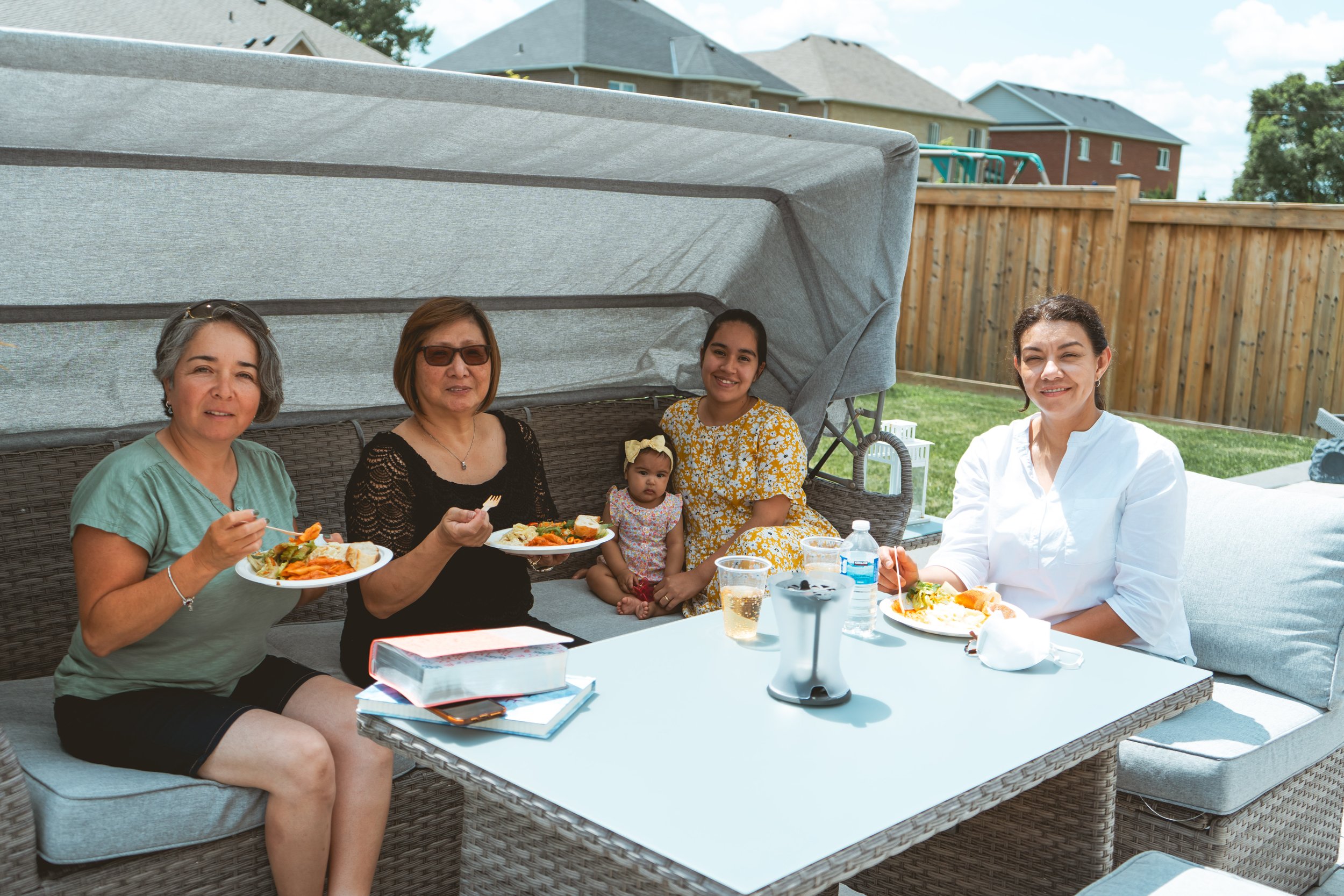 A group of four women and a baby sitting on outdoor patio furniture eating a meal. They are in a backyard with a wooden fence and houses visible in the background.