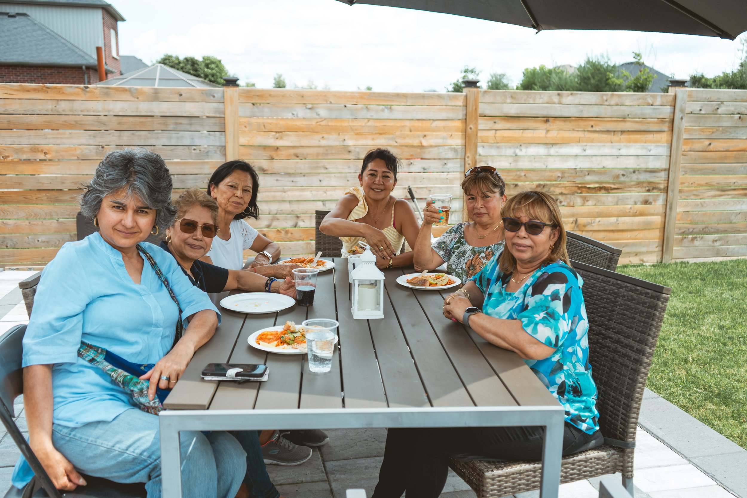 A group of six women sitting at an outdoor dining table, enjoying a meal and drinks under a large umbrella in a backyard. They are smiling and posing for the camera.