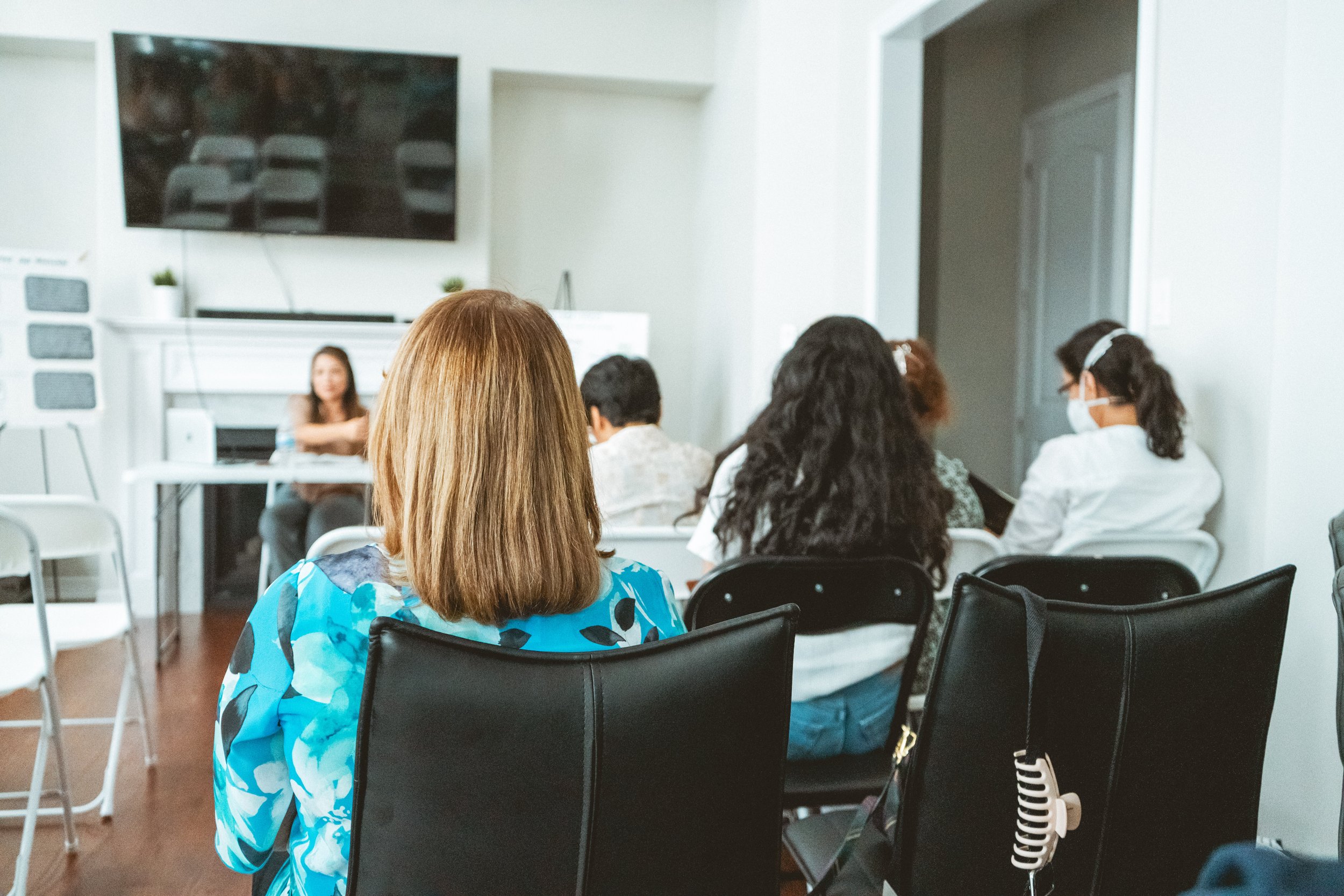 People seated in a meeting room, focus on woman's back with floral shirt, TV on wall, speaker at the front, attendees in casual attire.