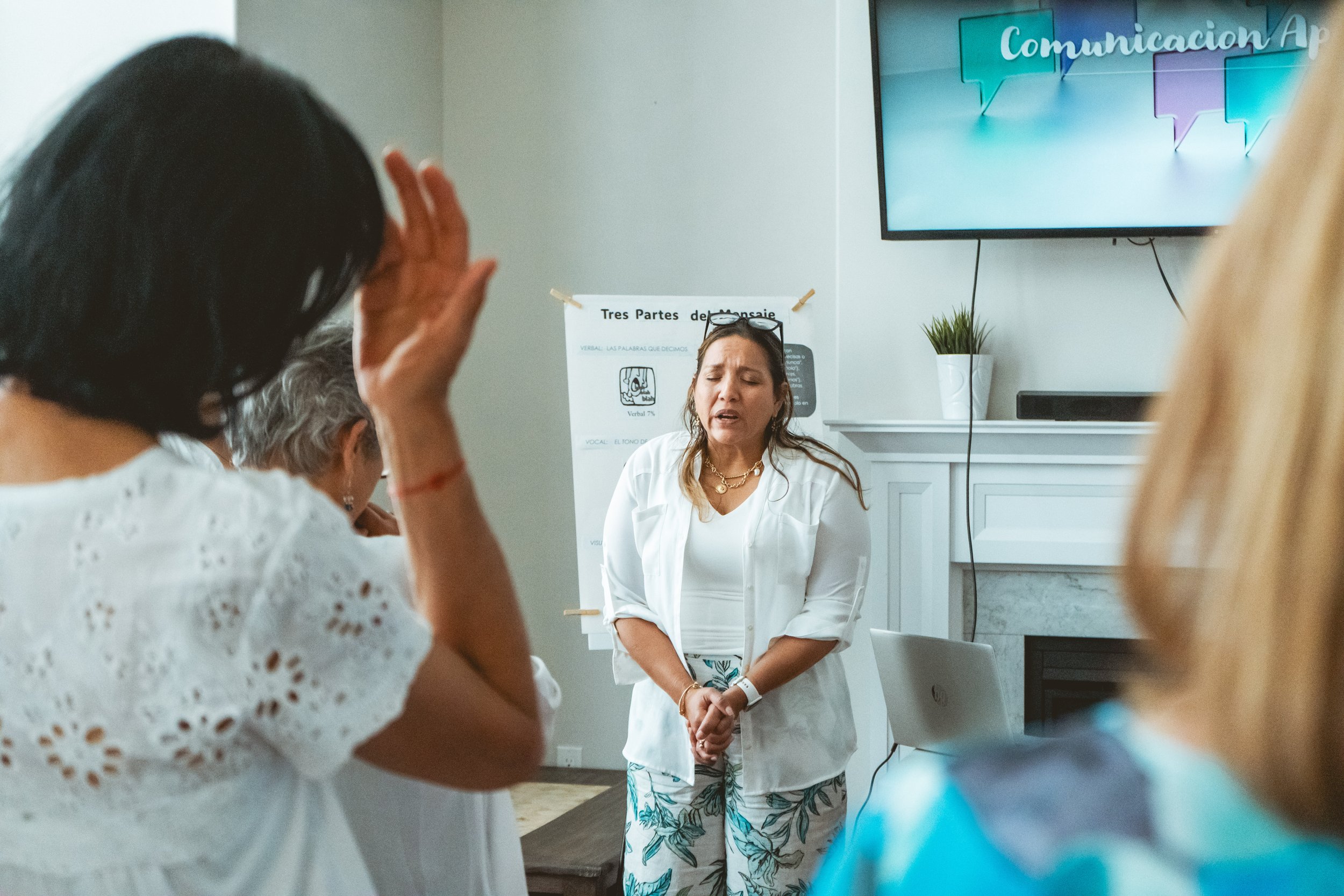 A woman in white standing in a room with people, leading a discussion or workshop. A poster with Spanish text and a TV screen are in the background.