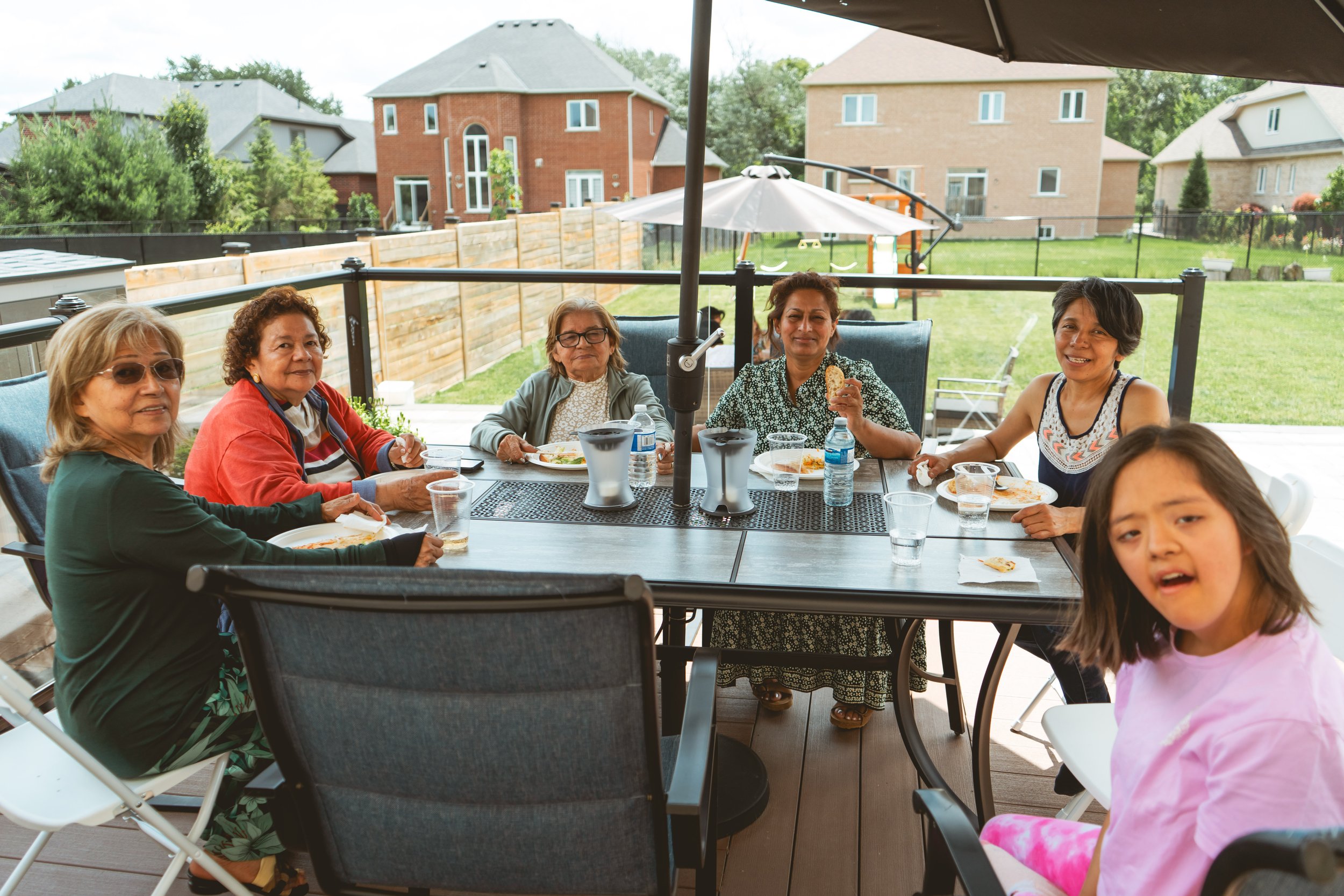 A group of people sitting around an outdoor table, enjoying a meal on a patio. There are houses and a lawn in the background.