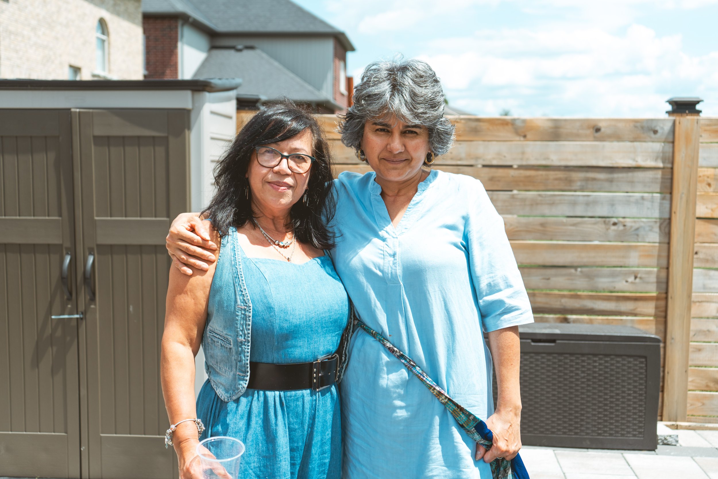 Two women smiling outdoors, one in a denim outfit and glasses, the other in a light blue dress.