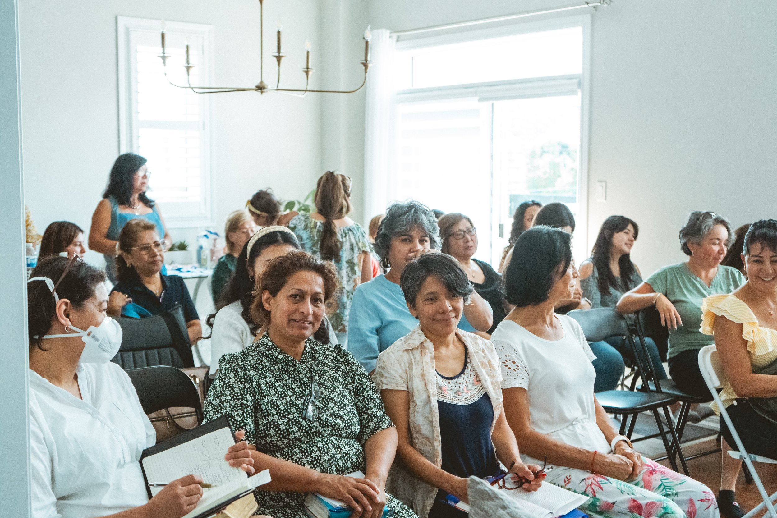 Group of women sitting in a room, some smiling, others taking notes, in a casual meeting setting.