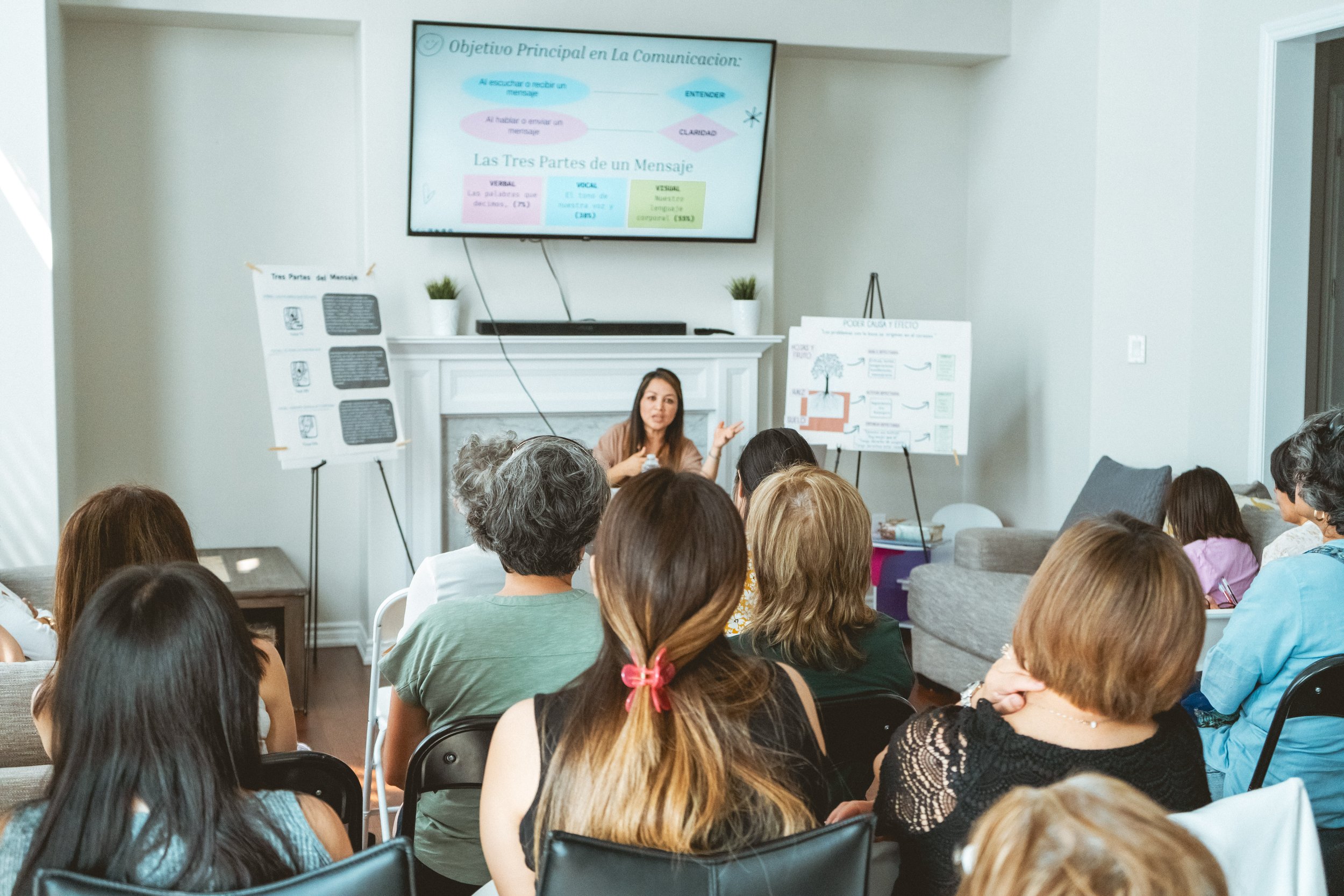 A woman giving a presentation to a seated audience in a living room. A large screen displays a slide in Spanish, and easel boards with diagrams are positioned nearby. The attendees are focused on the speaker.