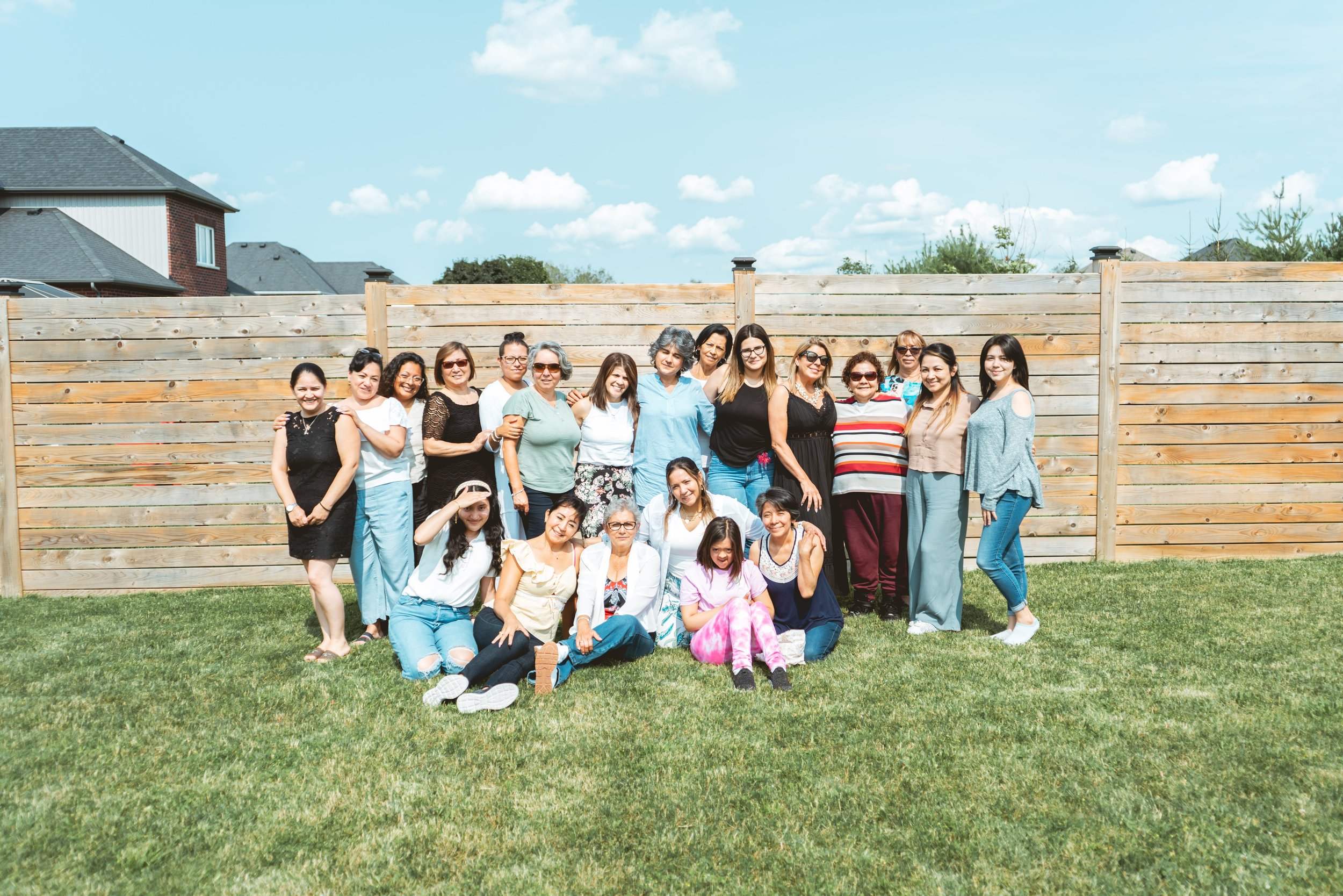 A group of women posing for a photo in a grassy backyard with a wooden fence and houses in the background. Some are standing while others are crouching or sitting on the grass. The sky is clear with a few clouds.