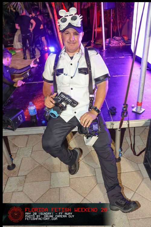 DC Photographer at Florida Fetish Weekend , standing on a stage with equipment, wearing a white shirt, with goggles on his head, holding camera gear, in an indoor event space with people in the background. Celebrating winning award that night 2025.