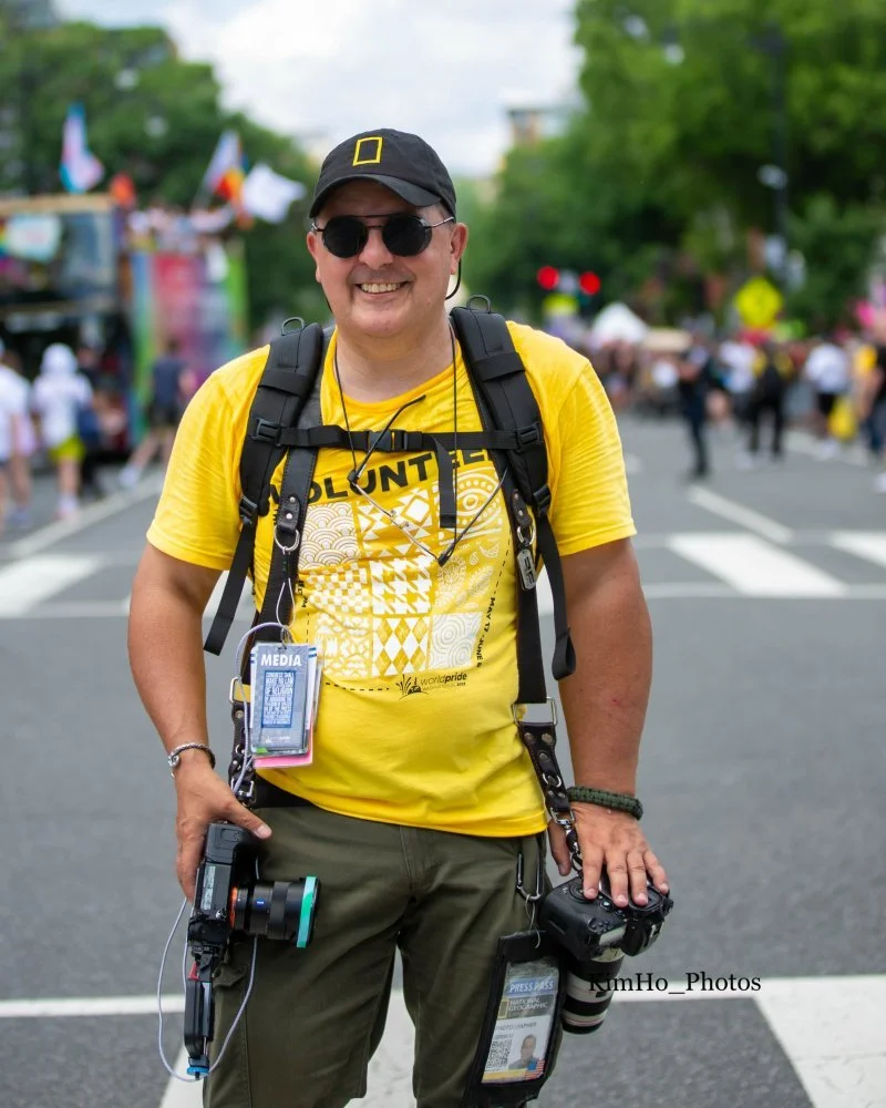 A smiling person wearing sunglasses, a black cap with a yellow logo, and a yellow t-shirt standing on a street at a parade or event. They have a black backpack and media badge, and are holding cameras and recording equipment. In the background, there are blurred people and flags, indicating a lively outdoor gathering.