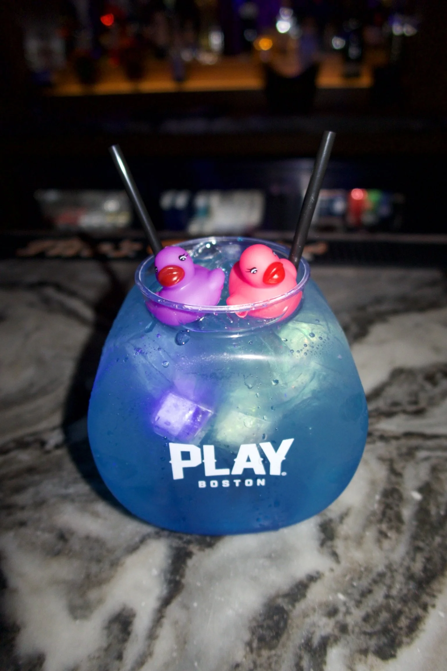 A blue cocktail in a rounded glass with 'PLAY BOSTON' written on it, with ice cubes inside, topped with two small rubber duck toys and two black straws, set on a marble surface at night.