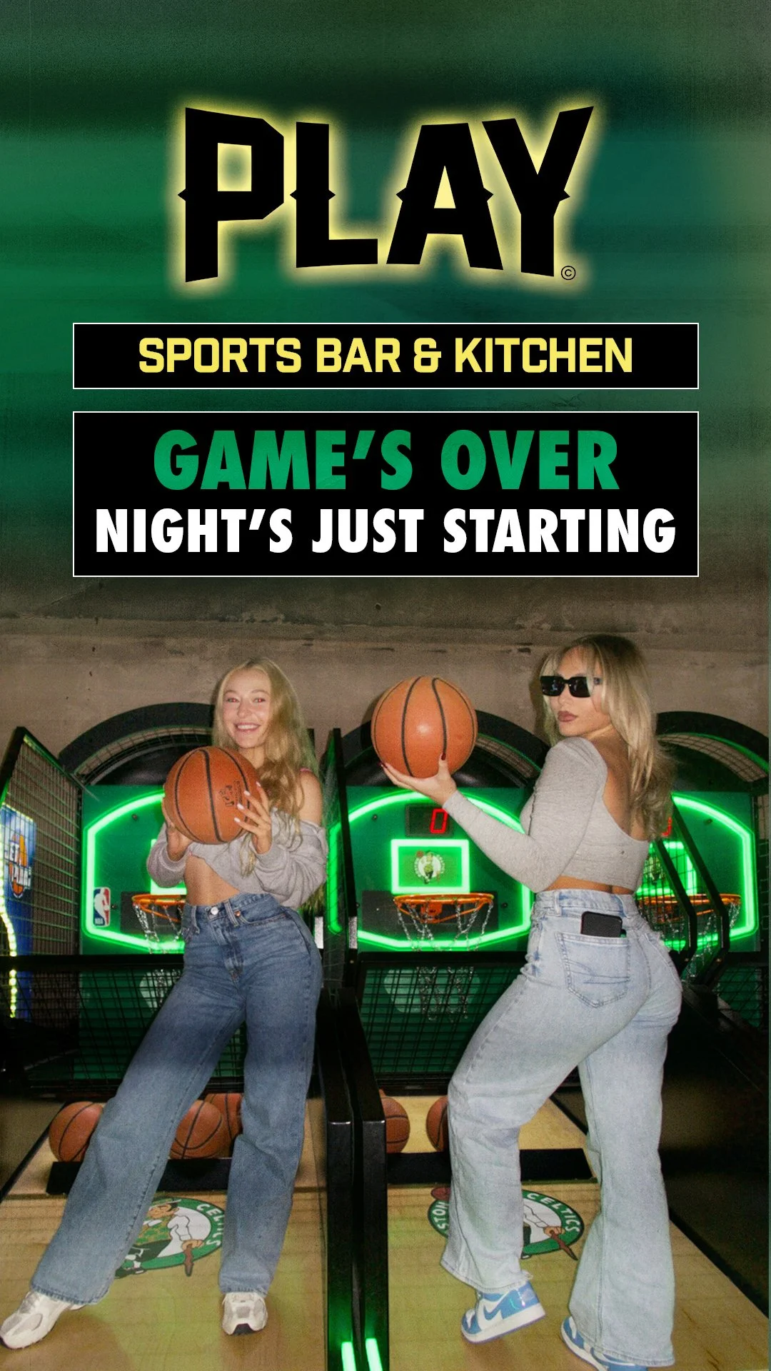 Two women playing basketball arcade game at a sports bar with neon lights, with a sign above indicating the bar is open and the game is just starting for another night.