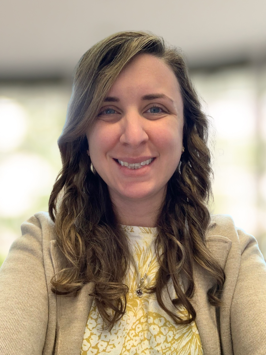 A woman with long, wavy brown hair and blue eyes smiling at the camera, wearing a beige jacket over a yellow and white patterned top, with a blurred bright background.