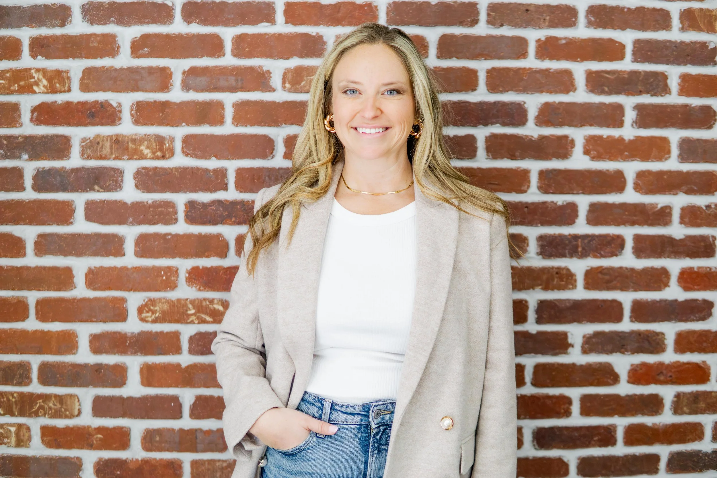 A woman with blonde hair and blue eyes smiling, standing in front of a red brick wall, wearing a beige blazer, white top, and blue jeans.