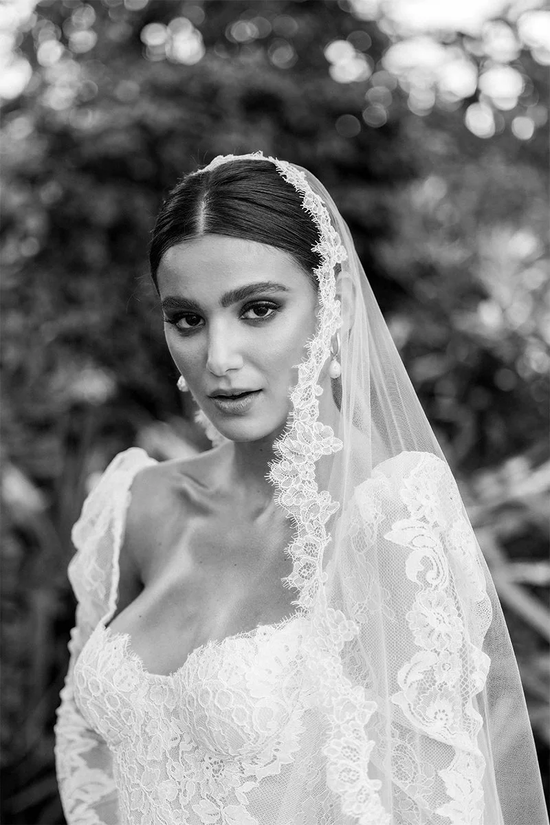 Bride in lace wedding dress and veil in a garden setting, black and white photo.