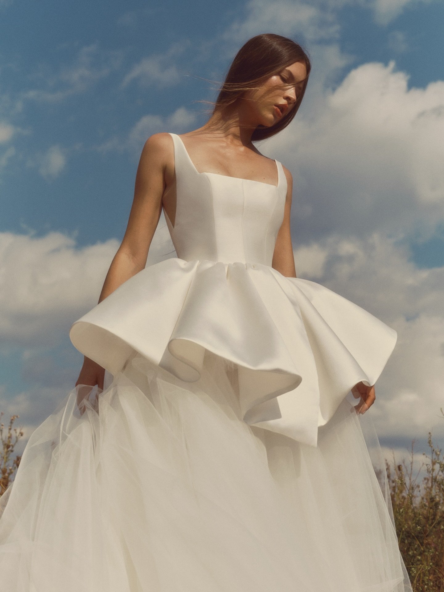 Meet RIPPLE 🤍 Sculpted ballgown with a modern square neckline and delicate straps that dip into a V at the side. The seamed bodice unfolds into a layered peplum that splits into soft points at the front and back, floating over an airy tulle skirt. A