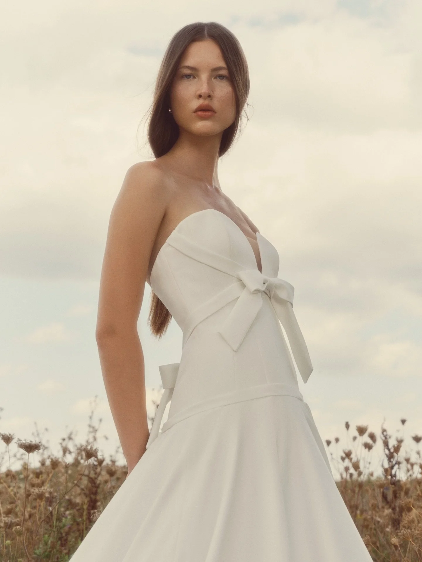 PEARL ✨ Strapless ballgown with a sculpted sweetheart, elongated bodice wrapped in soft bias bands that drape and tie into delicate bows at the front and back. The full skirt sweeps with airy volume, while pearl buttons trace the back to the hem.

Ri