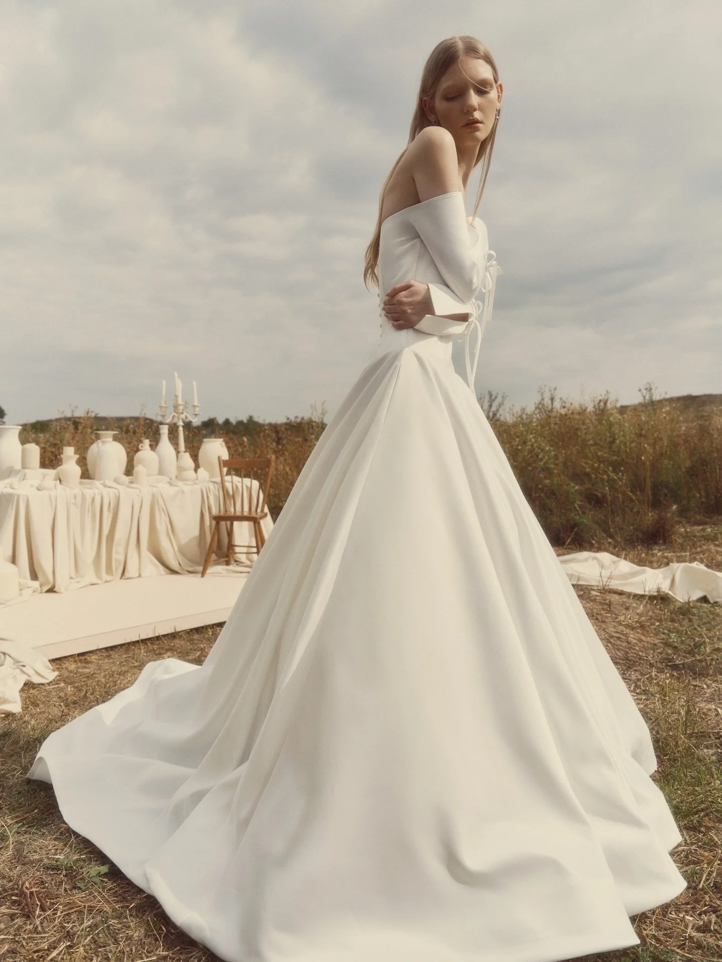 Off-the-shoulder ballgown with voluminous proportions and long tailored sleeves. The flared sleeves open with a subtle slit, tied with a ribbon bow. Finished with pockets and buttons tracing the back to the hem- poised, dramatic, and quietly regal.

