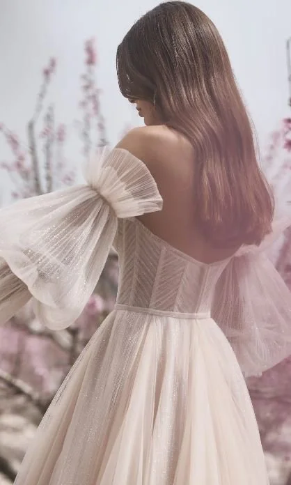 A woman with long brown hair wearing an elegant, off-the-shoulder, cream-colored dress with puffy, sheer sleeves.