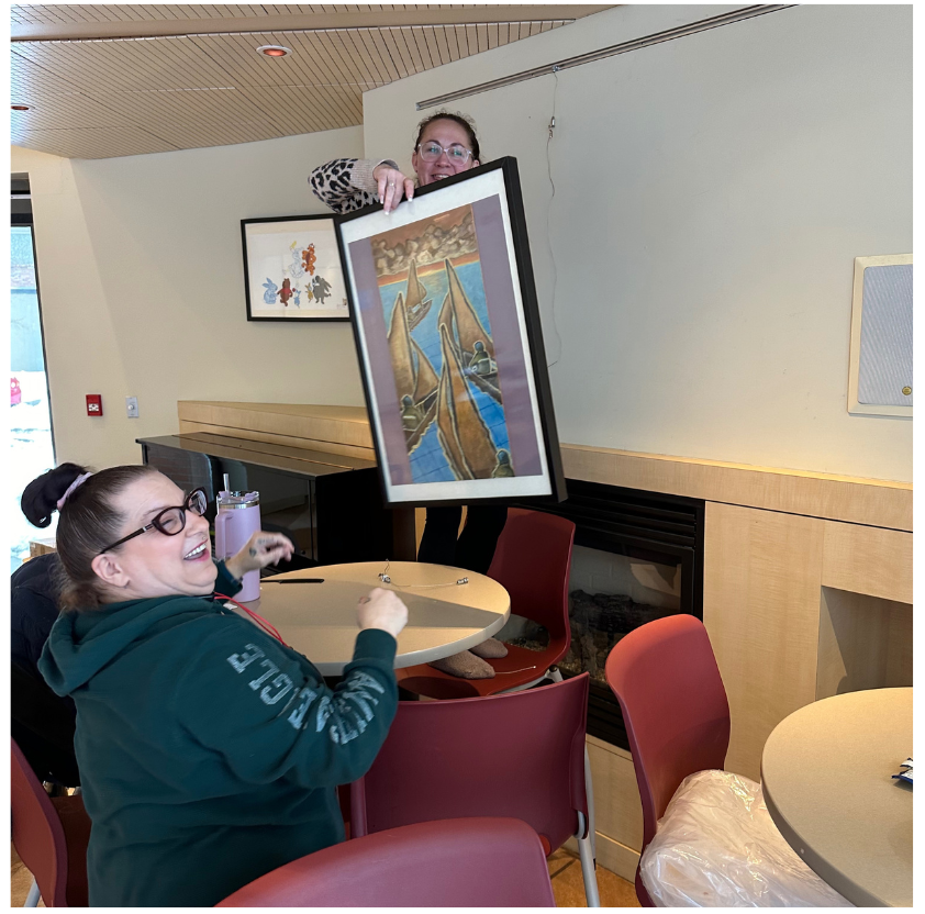 Two people are in action hanging a framed artwork while smiling