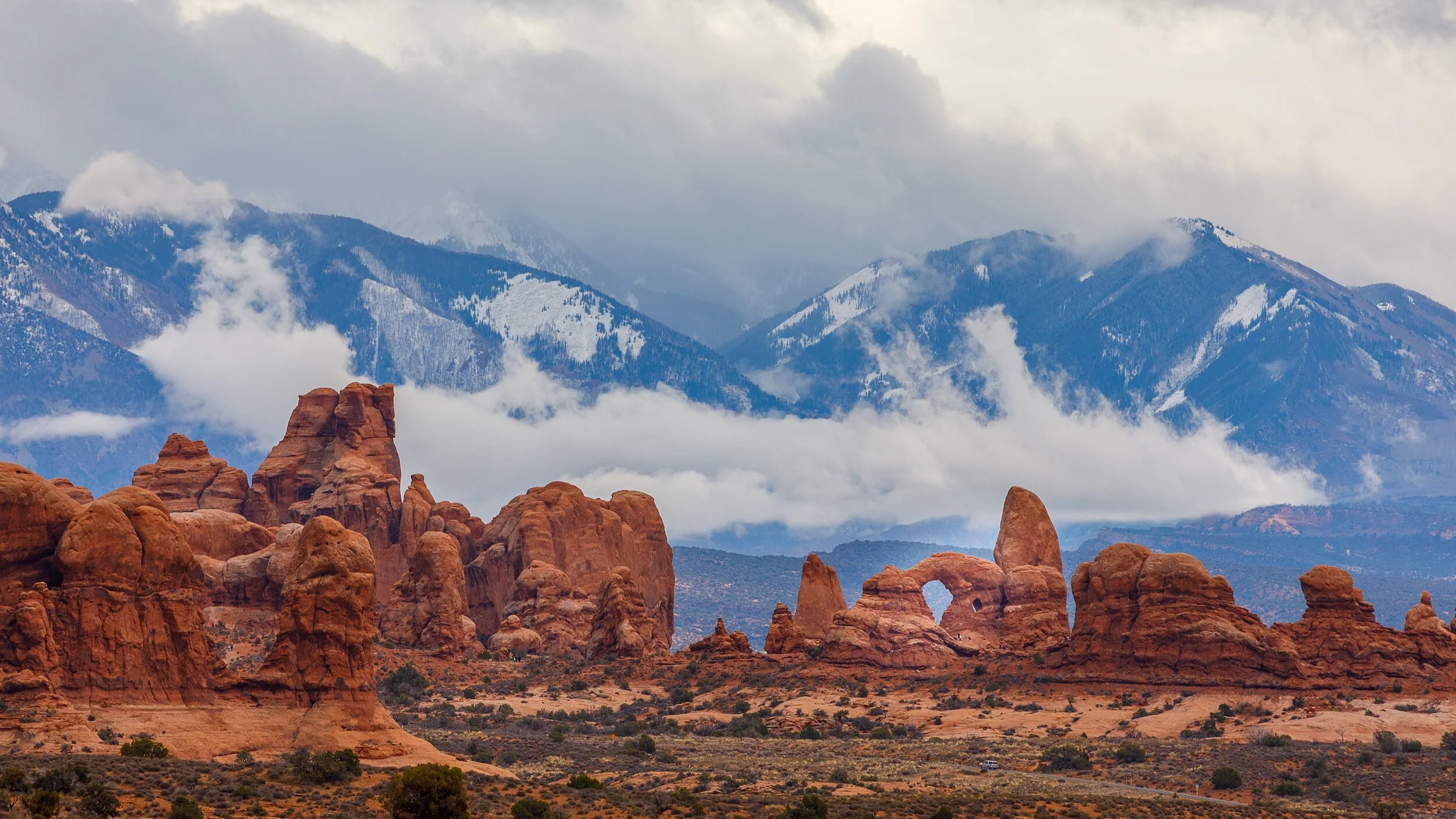 Red rock formations in a desert landscape with distant snow-capped mountains and cloudy sky