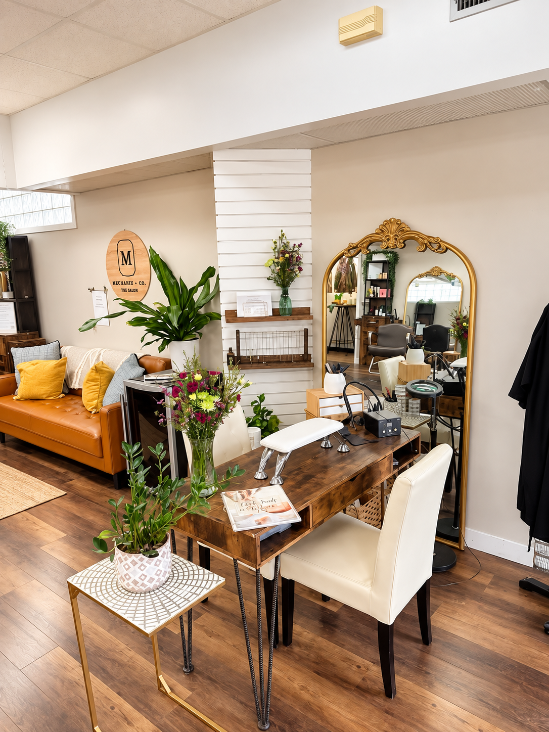 Interior of Mechanix + Co. salon in West St. Paul, with a gold-framed mirror, styling station, leather sofa, plants, and polished wood floors.