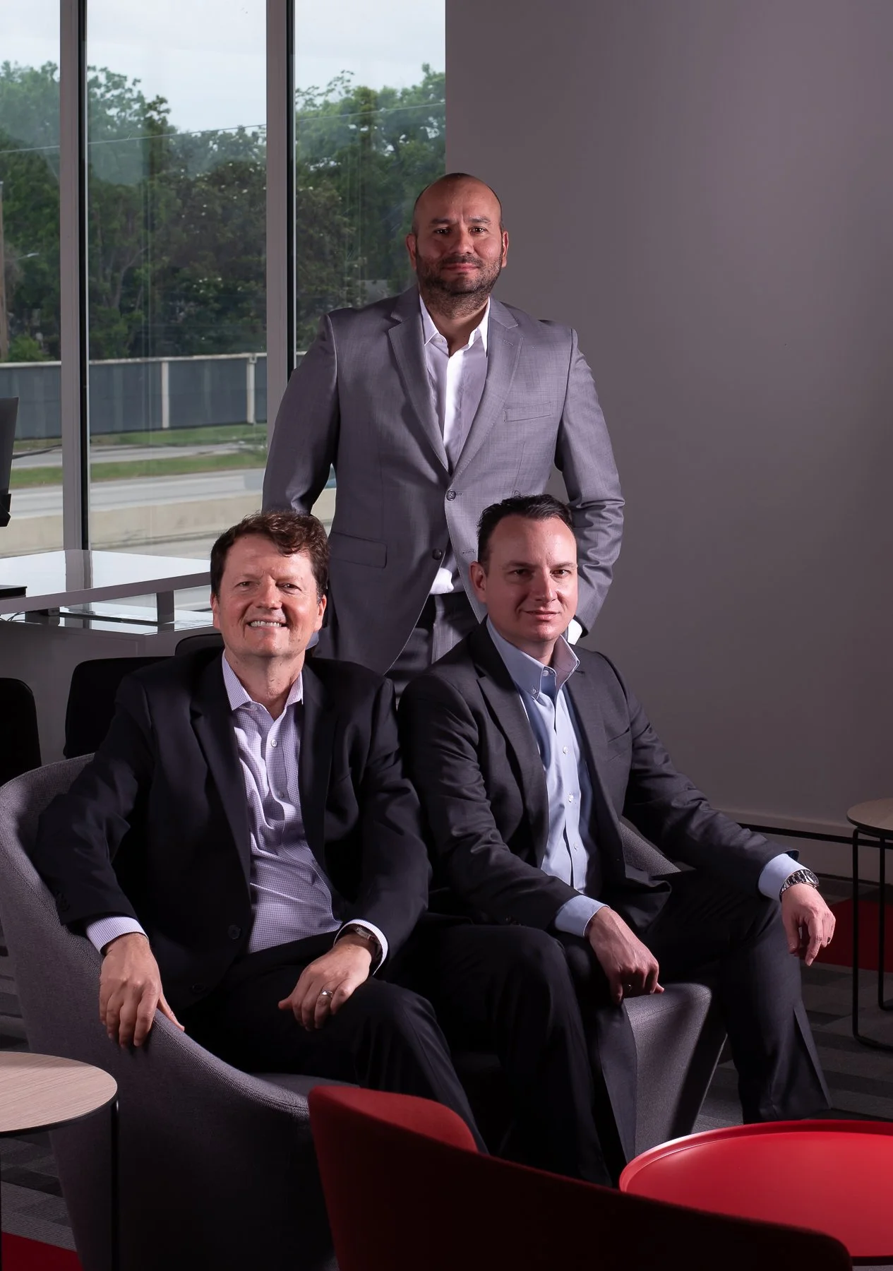 Three business professionals in suits posing in an office with large windows showing trees outside.