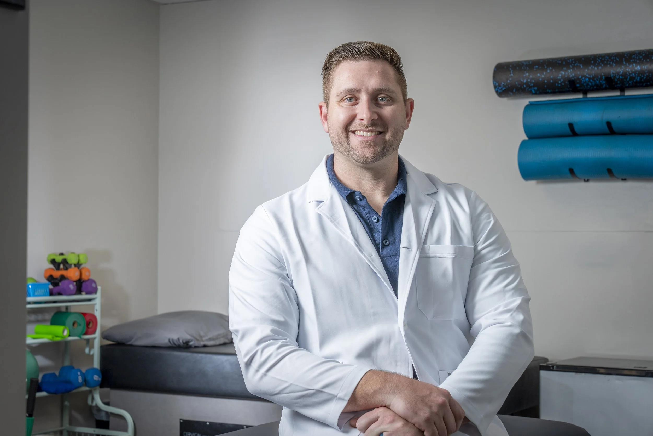 A smiling male doctor in a white coat standing in a medical office or clinic, with exercise mats and colorful exercise equipment in the background.