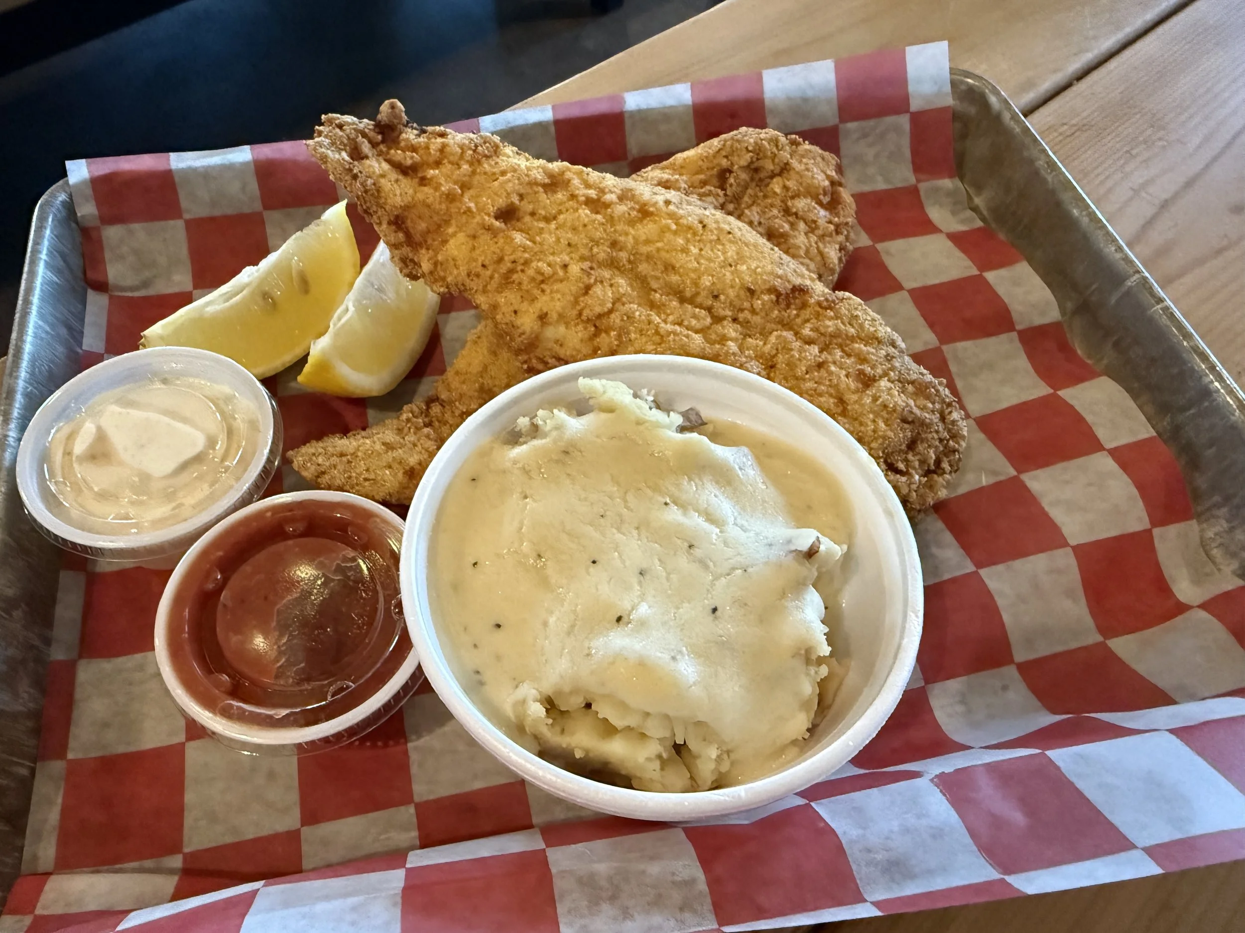 Fried fish fillets with lemon wedges, mashed potatoes with gravy, and dipping sauces on a checkered paper-lined metal tray.