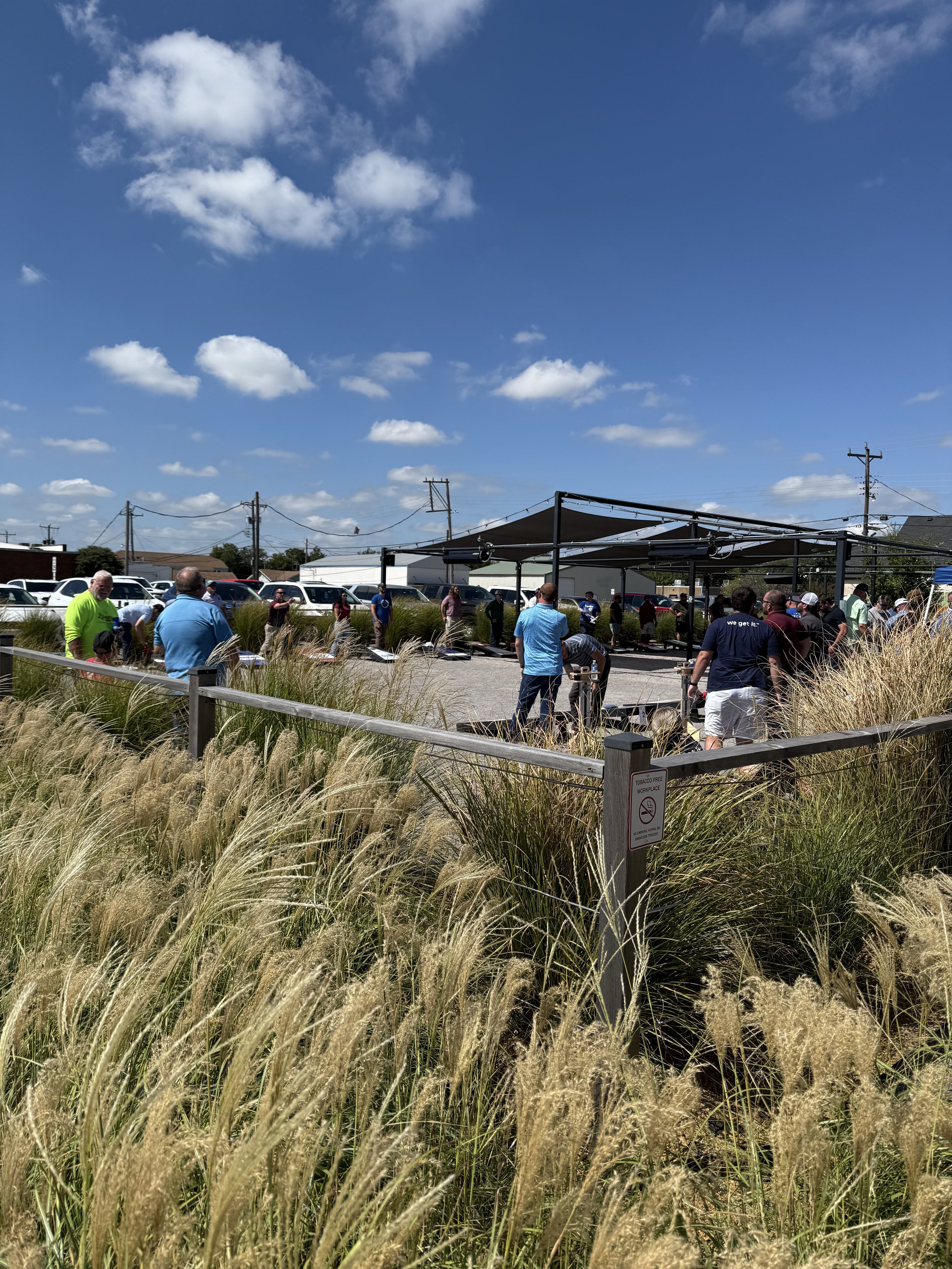 People gathered outside under a shaded structure on a sunny day, surrounded by tall grasses with a clear blue sky and scattered clouds overhead.