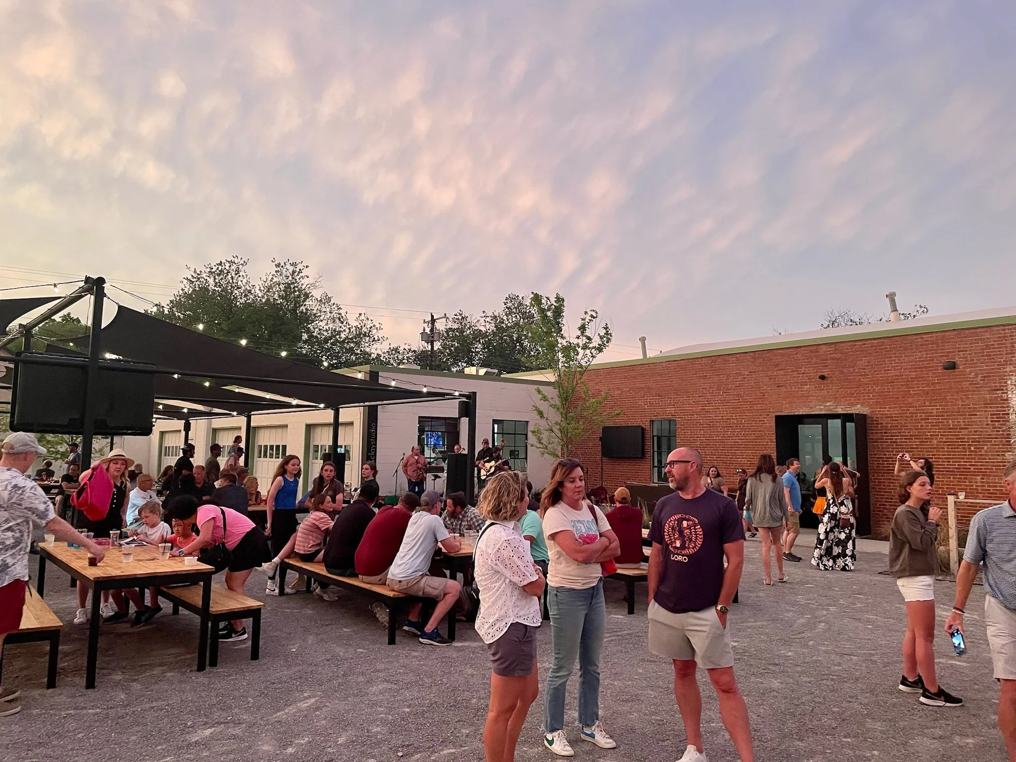 People gathered outdoors at a social event, some standing and talking, others sitting at picnic-style tables. A live band is playing on a stage with string lights, and the setting is next to brick and white buildings during sunset with a colorful sky